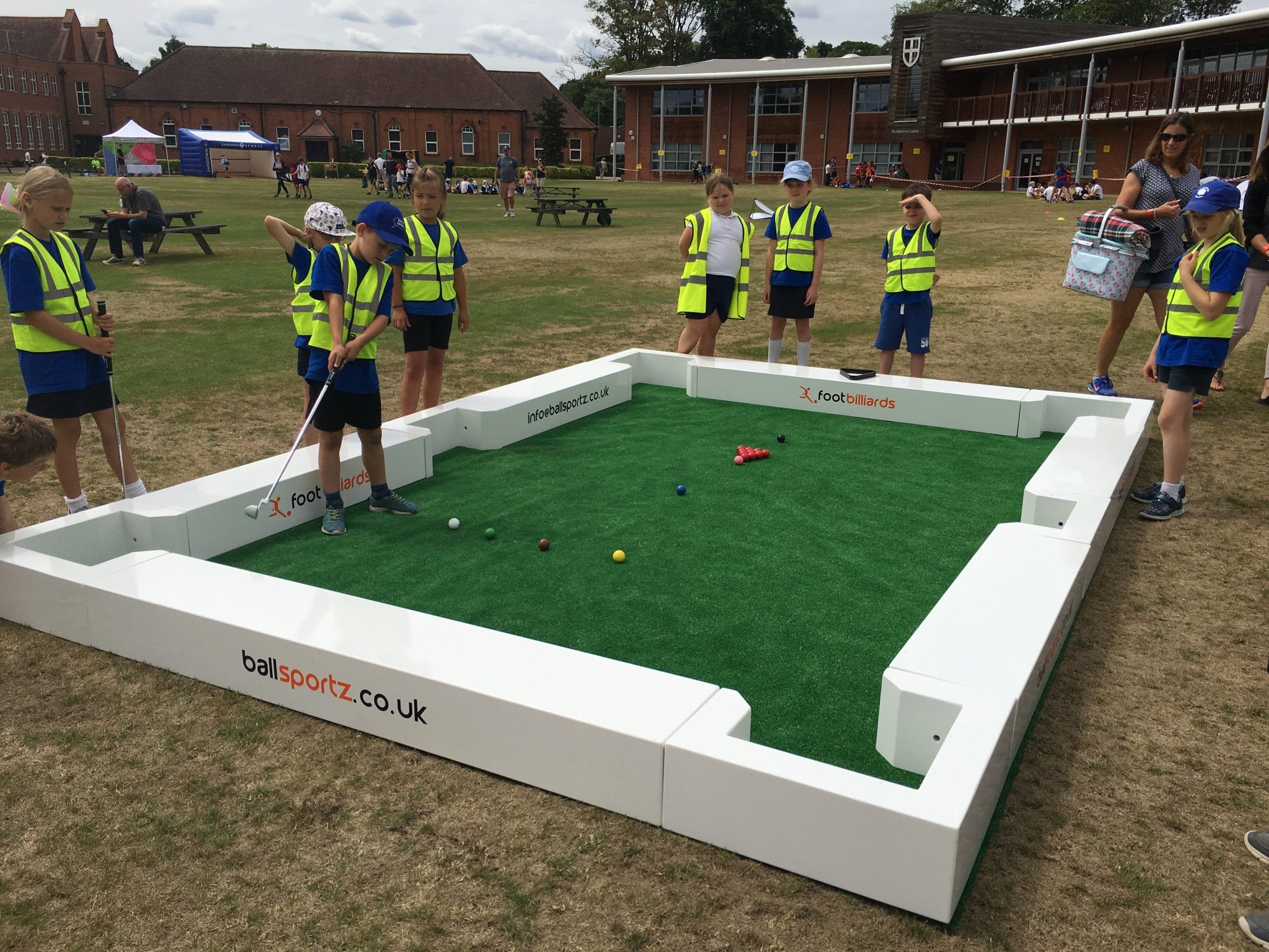 Children wearing high-visibility vests play a game of foot billiards on a green field set within a white-bordered arena.