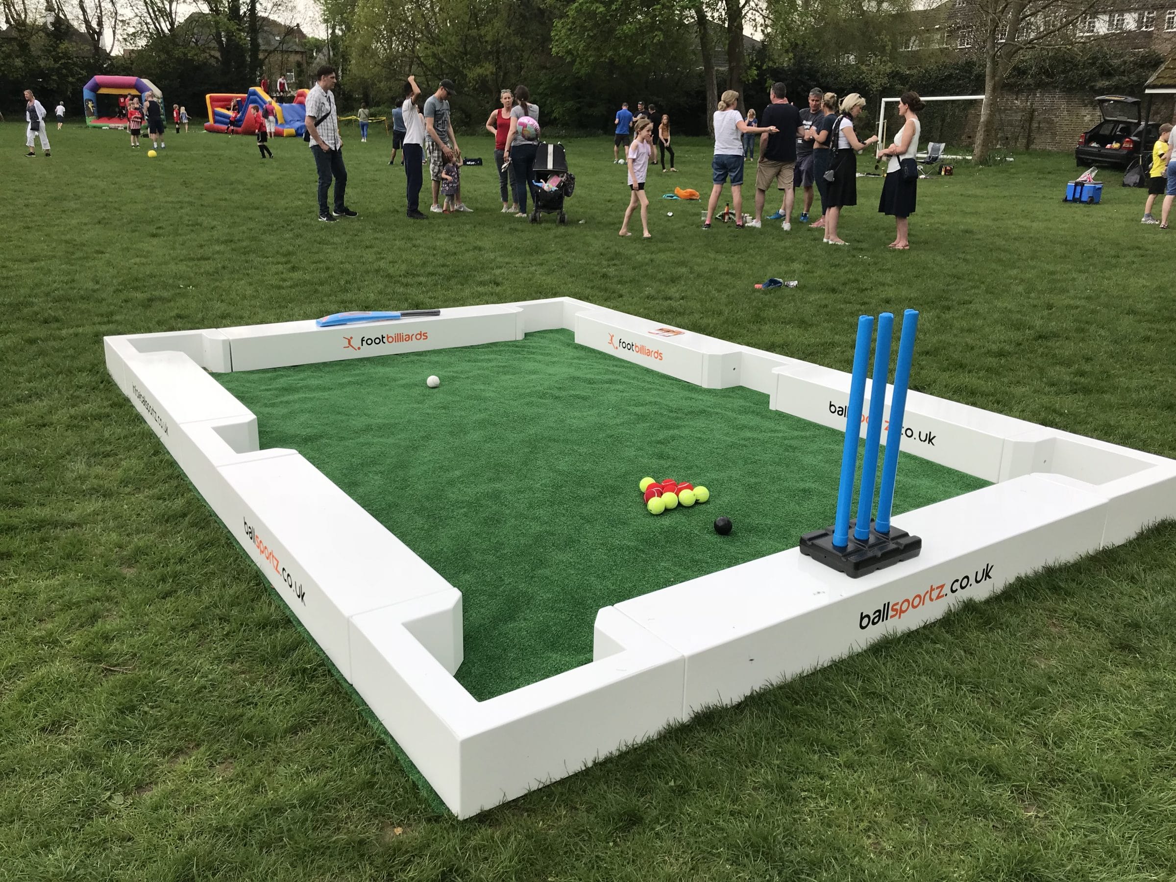 A mini cricket setup on grass with blue wickets and several colorful balls. People in the background are gathered around, some with children.