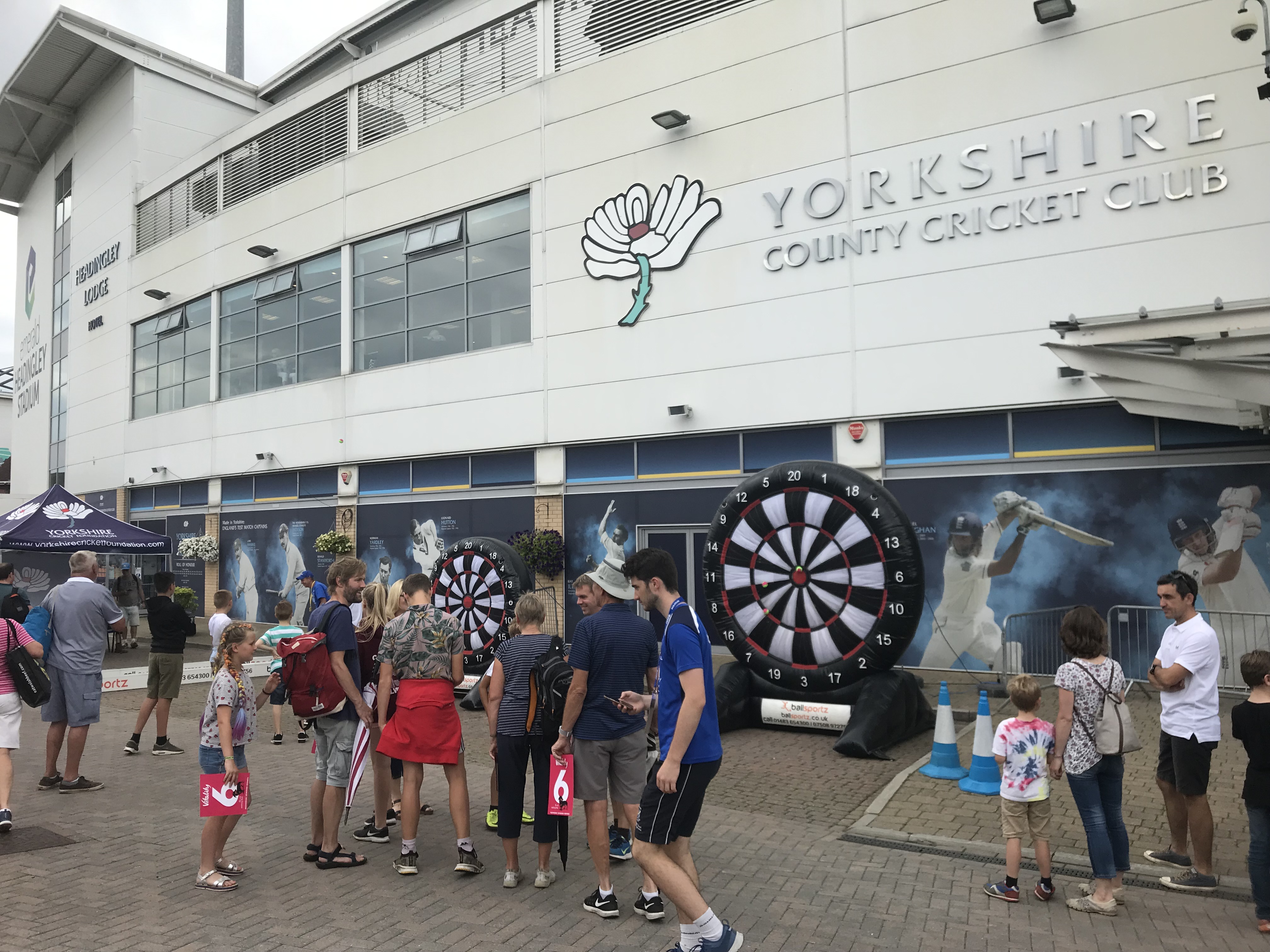 People gather outside Yorkshire County Cricket Club, with two large dartboards set up for an event.