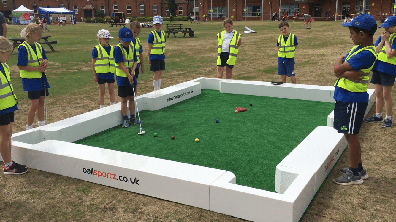 Children in safety vests and blue outfits play a mini-golf game on a grassy area, surrounded by a white fence.