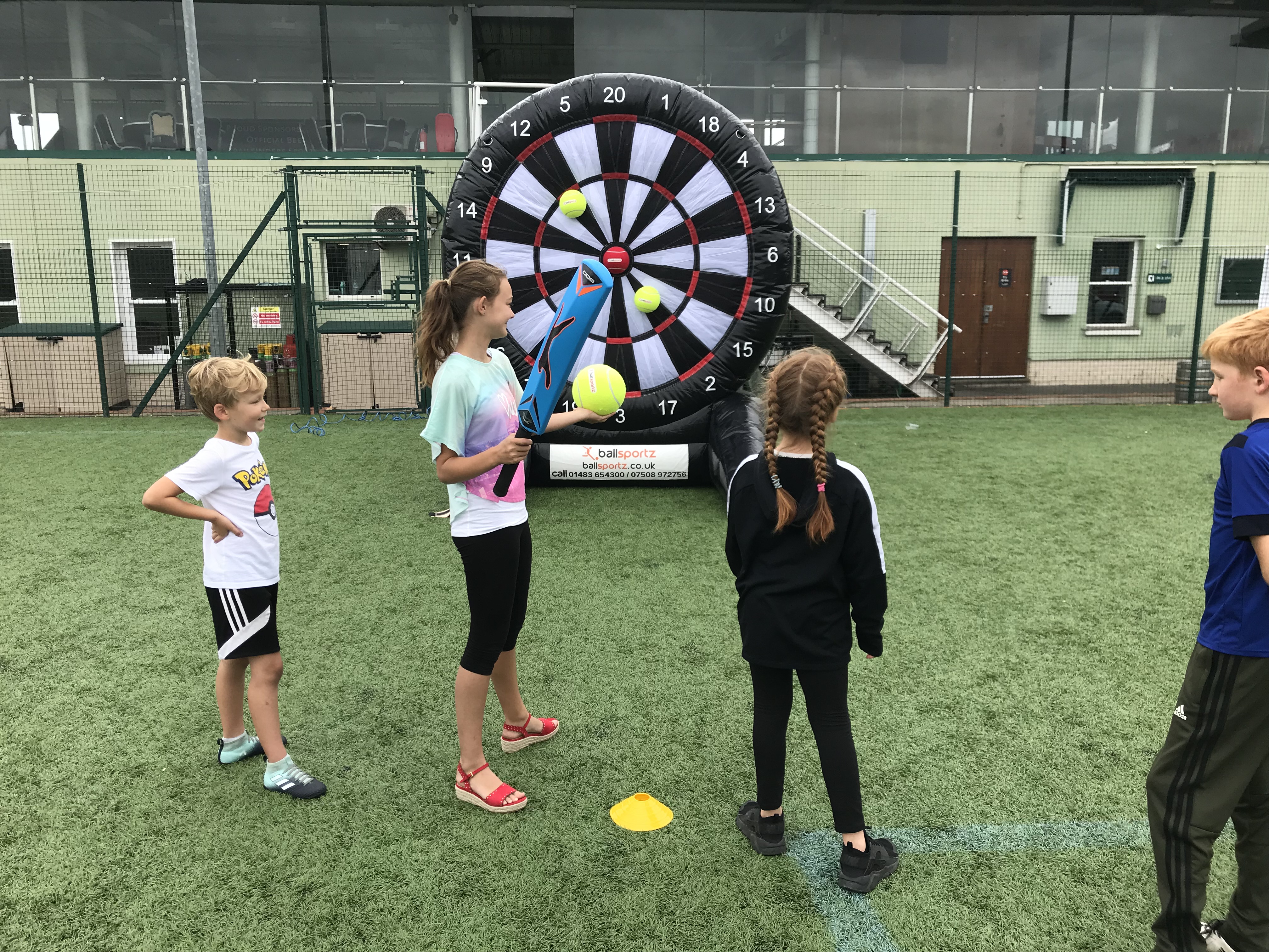 Children playing a game of soccer darts on a sports field, with a large inflatable dartboard in the background.