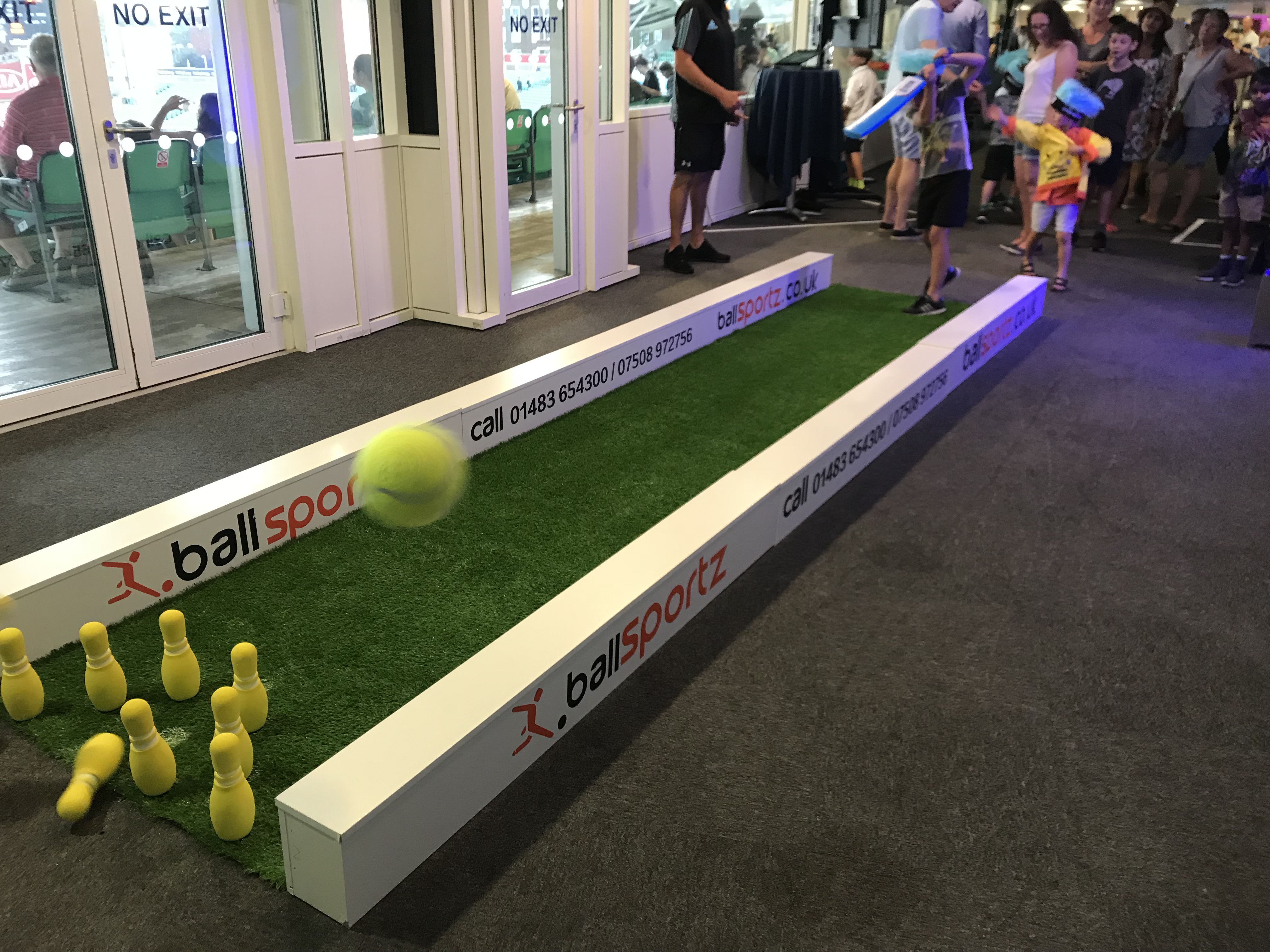 Children play a game of bowling using a tennis ball at an indoor venue, with sets of yellow pins on artificial grass.