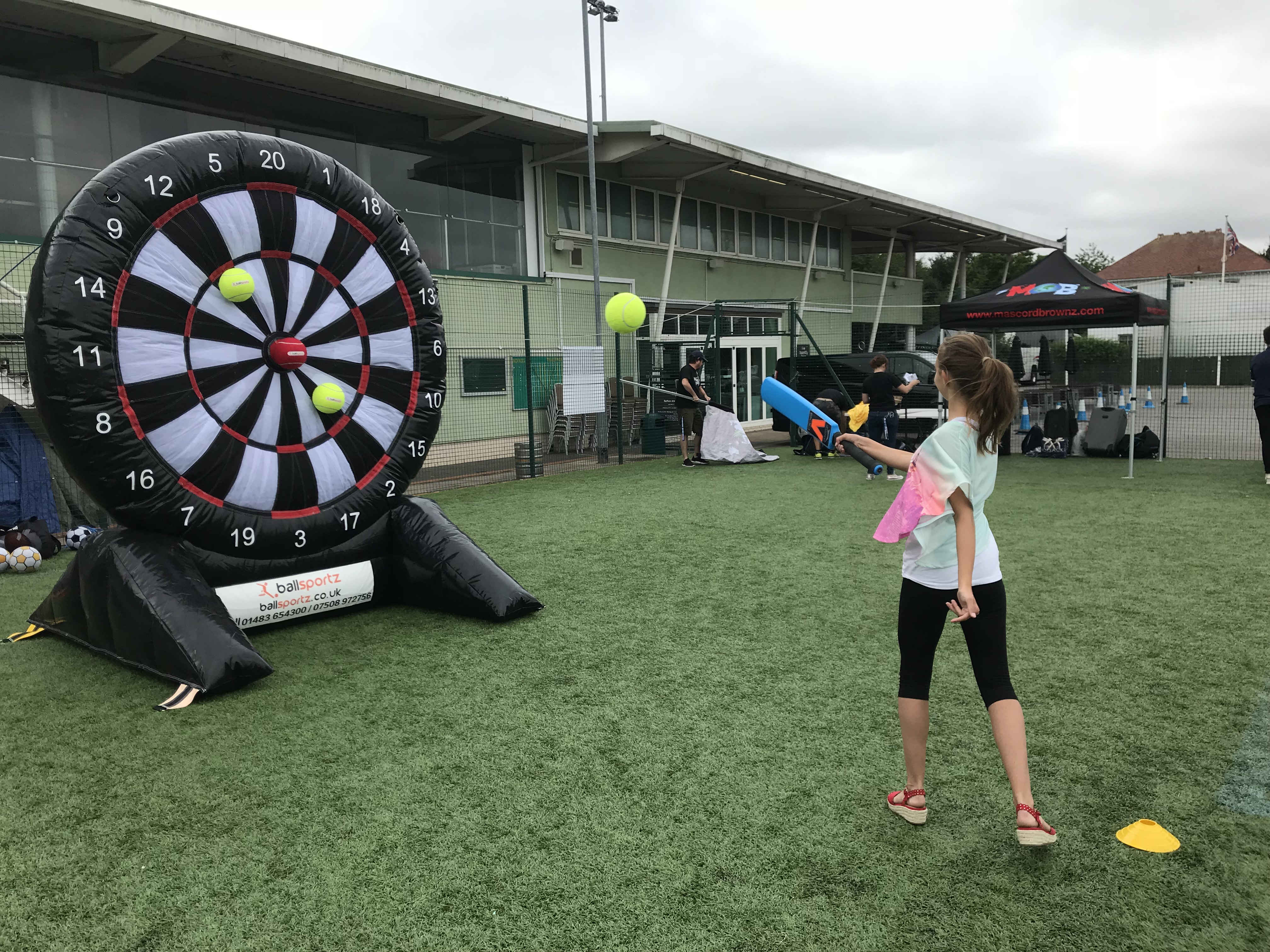 A person throws a ball at a large inflatable dartboard on a grassy field, with buildings and tents in the background.