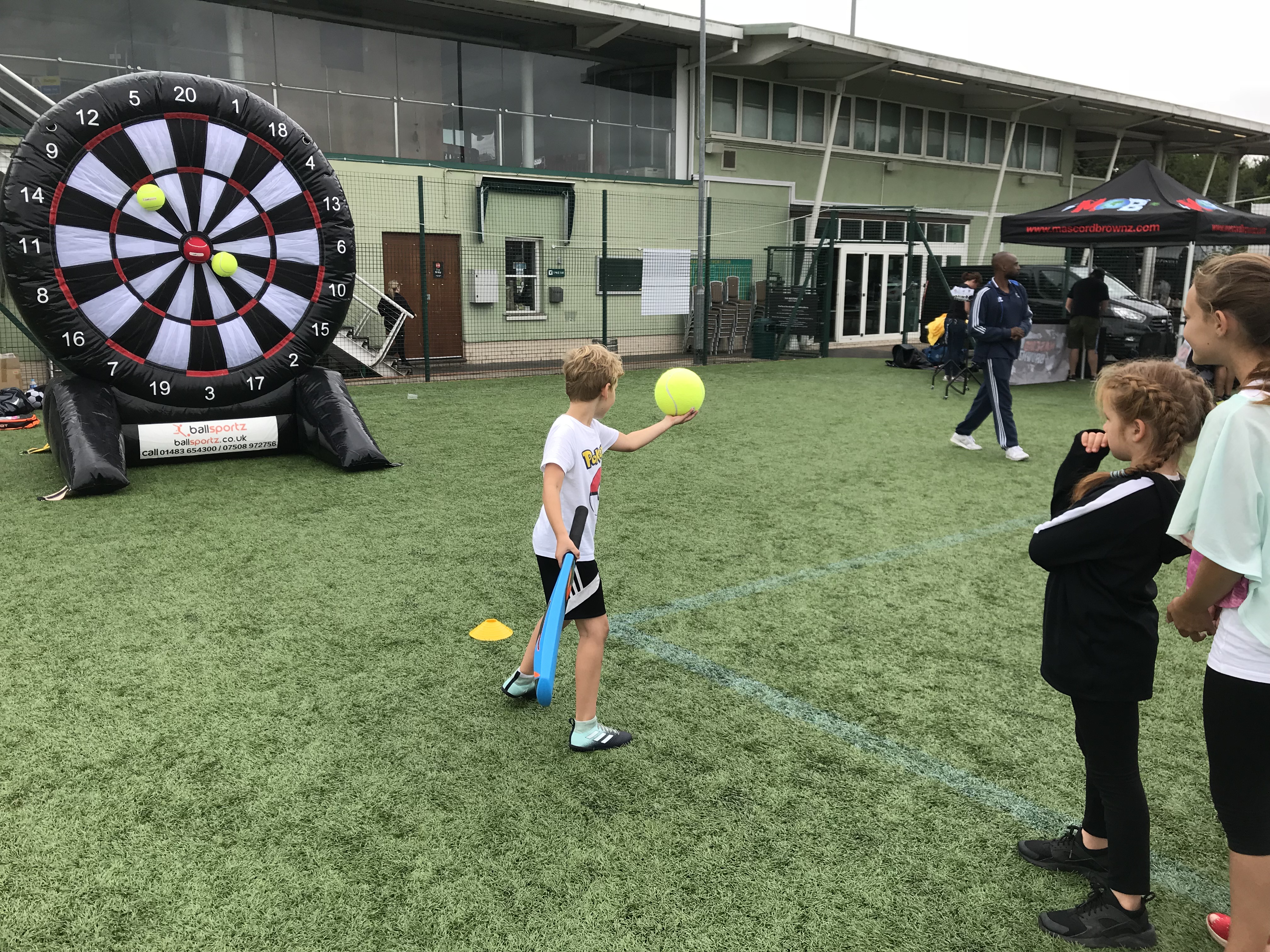 A child throws a large tennis ball at an inflatable dartboard on a sports field, as two others watch.
