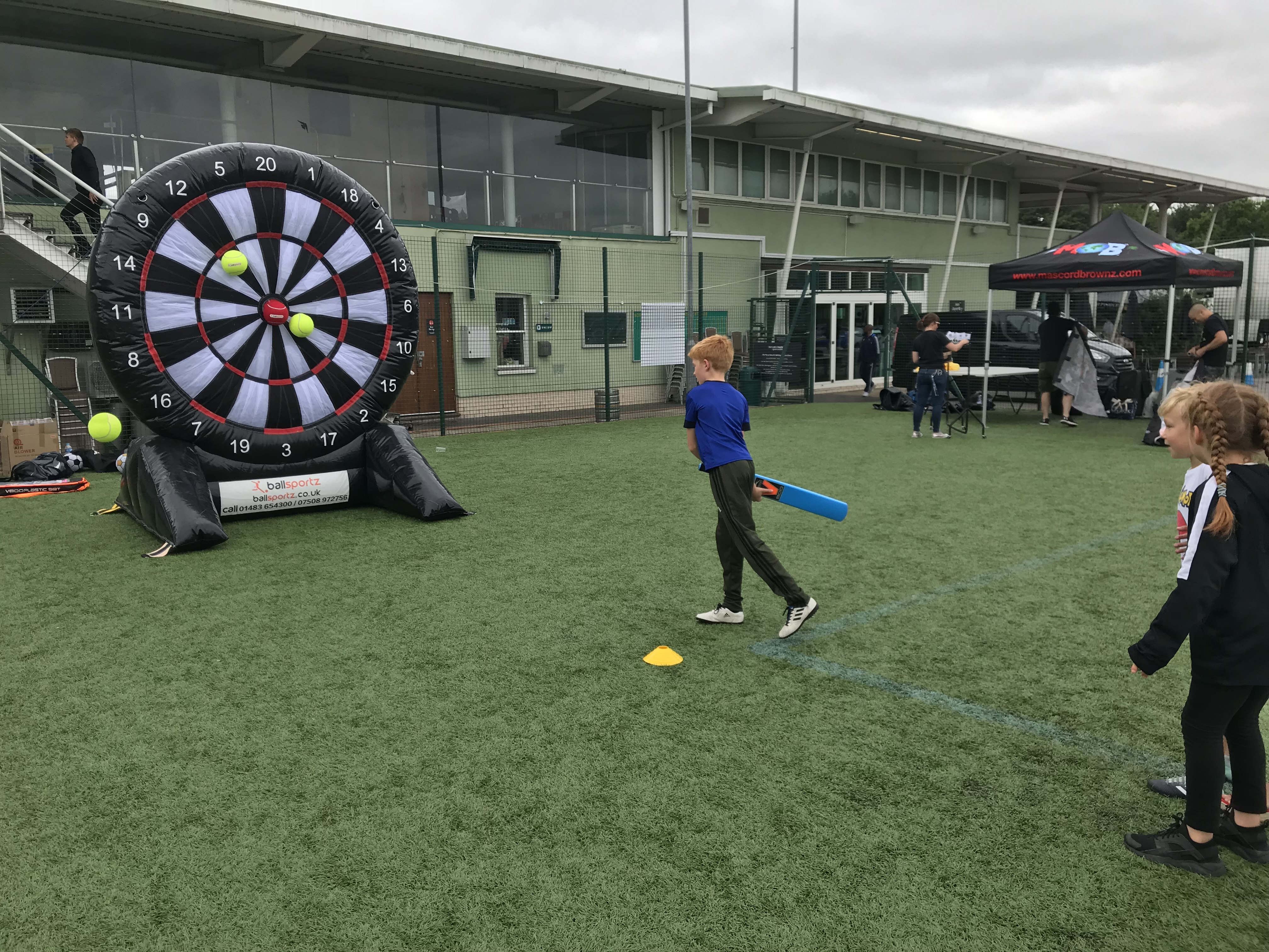 Children play a game by aiming balls at a giant inflatable dartboard on a grassy sports field.