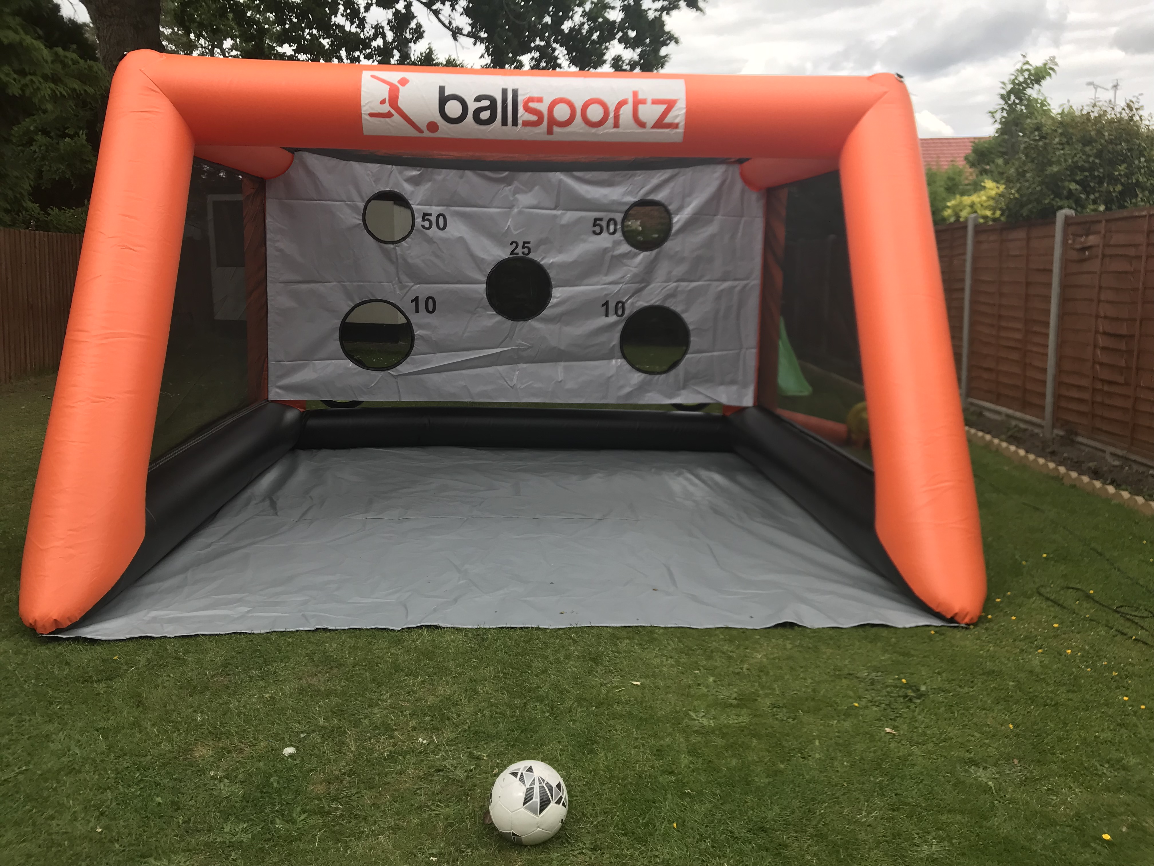 Orange inflatable soccer goal with target holes and point values, labeled "ballsportz," set up on grass with a soccer ball in front.