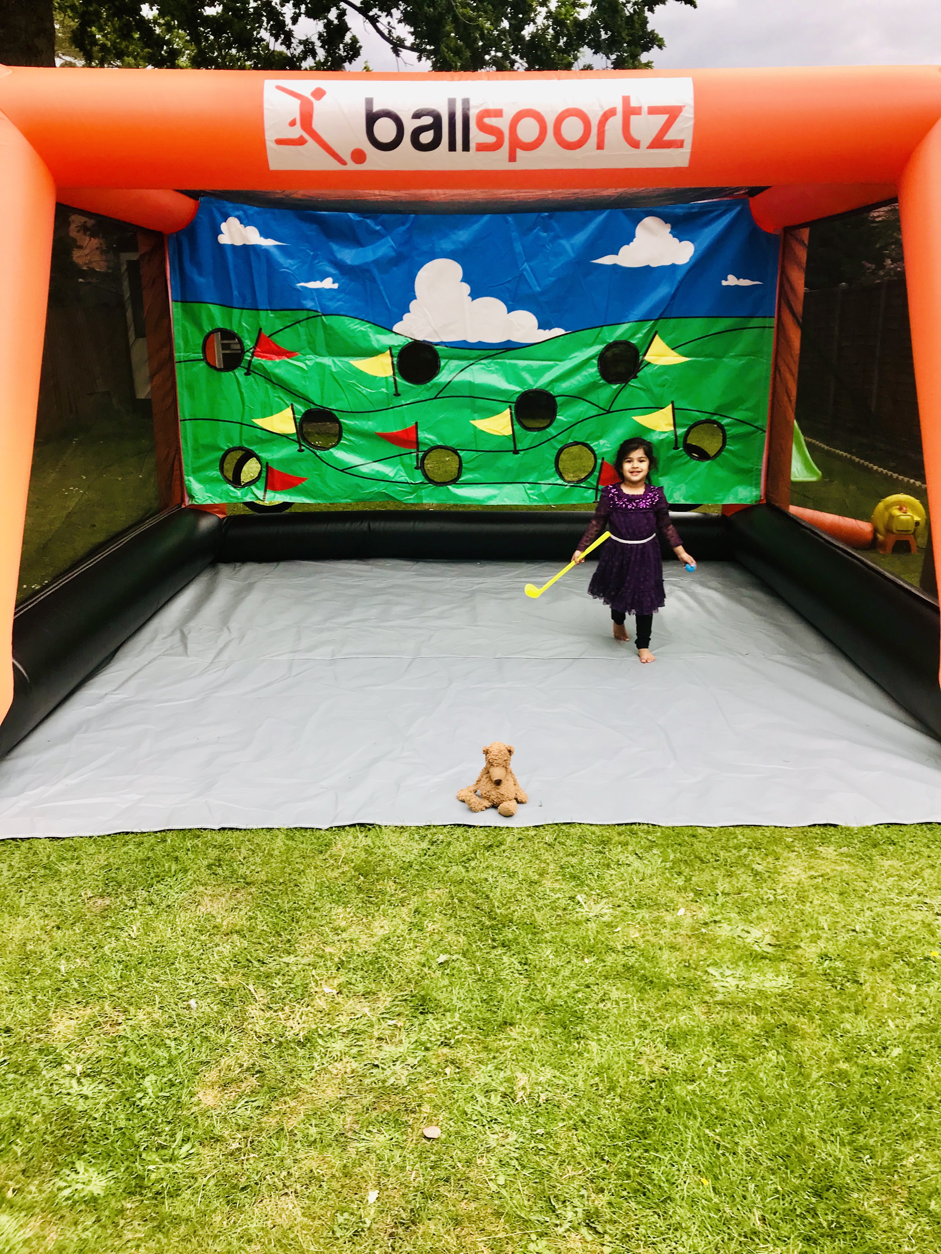 A child with a plastic golf club stands in front of an inflatable target backdrop labeled "Ballsportz." A teddy bear sits on the grass.