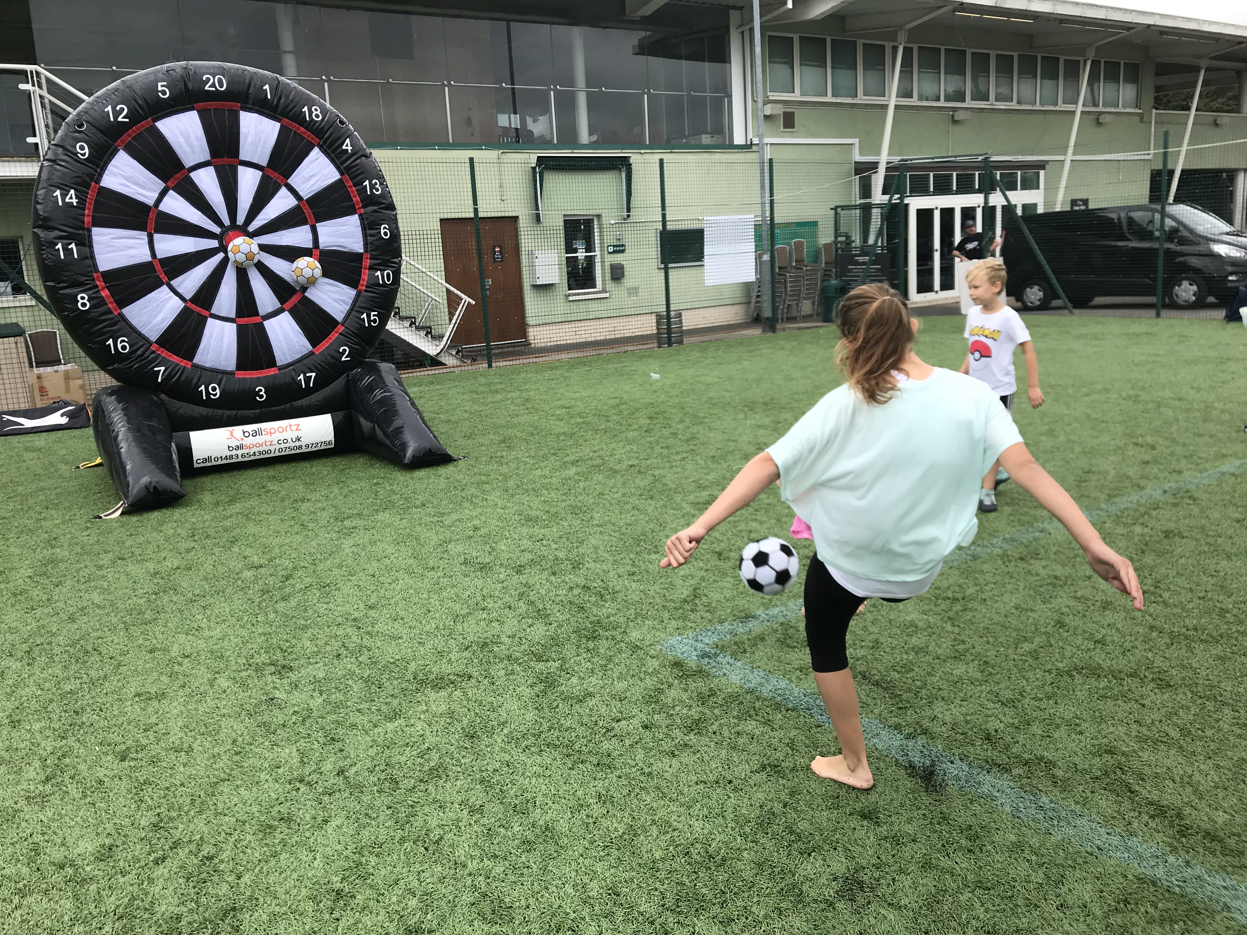 A person kicks a soccer ball towards an inflatable dartboard on a grassy field, while a child watches.