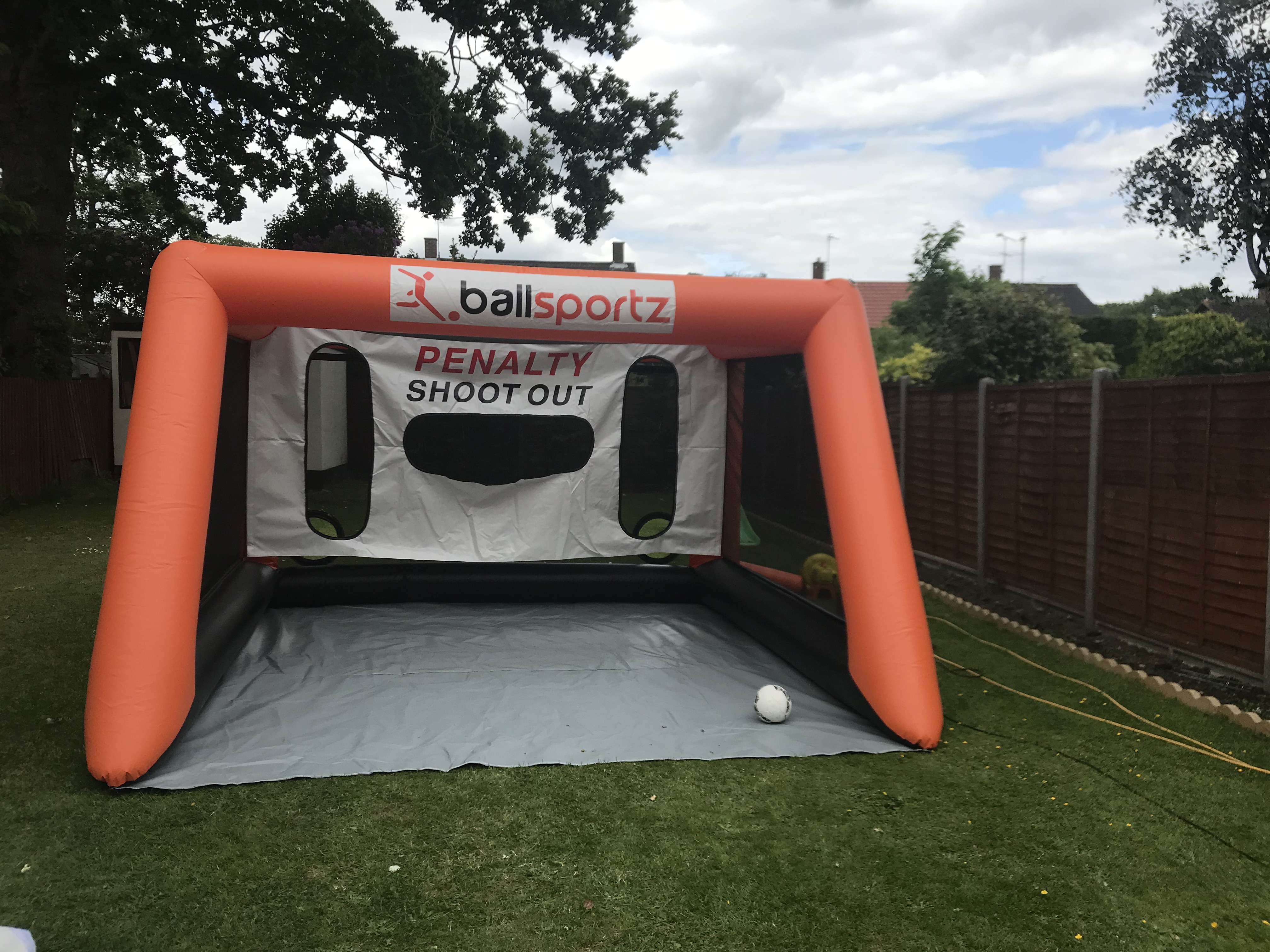 Inflatable penalty shootout game with an orange frame and a soccer ball on the ground in a backyard setting.