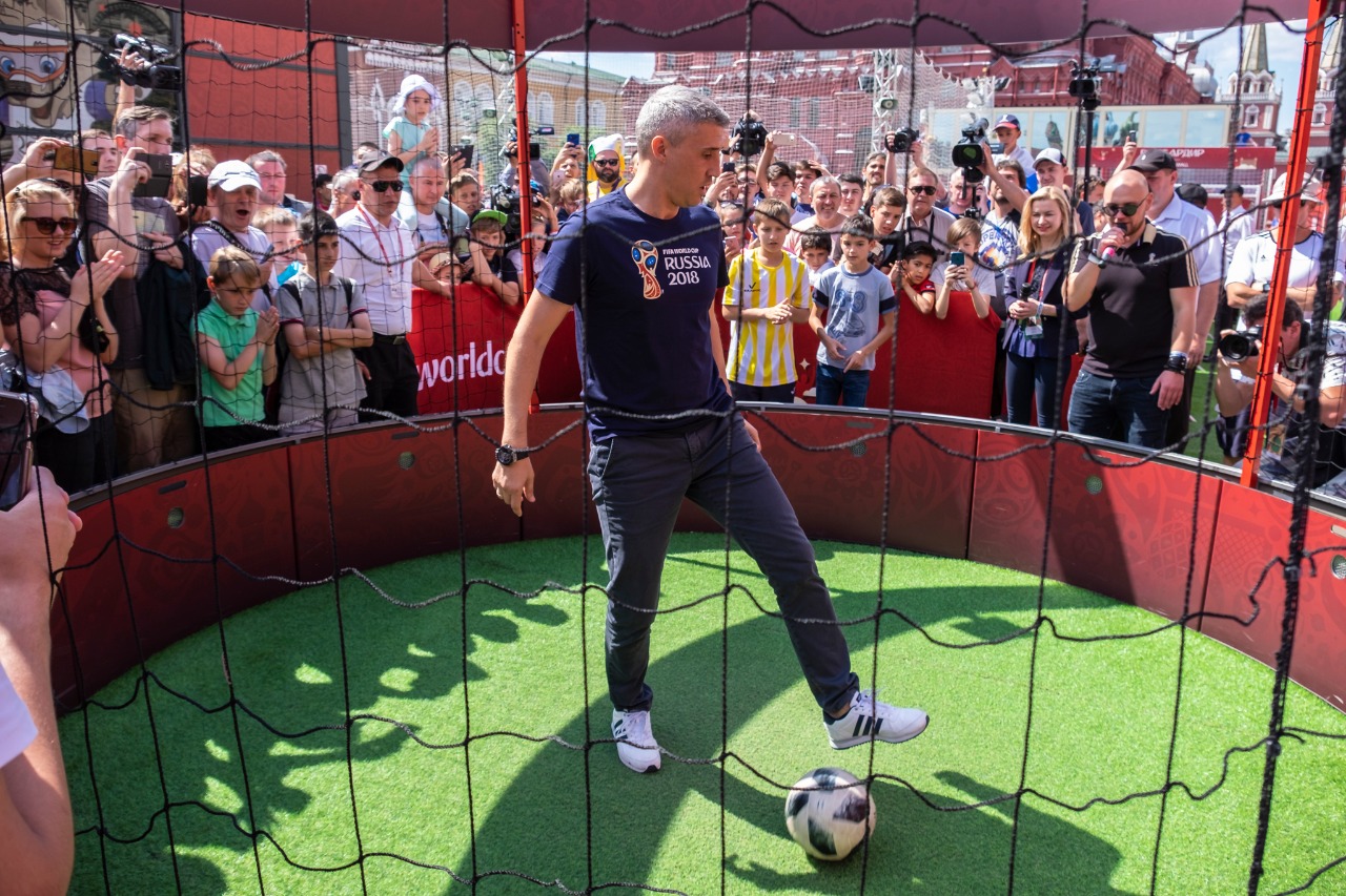 A man in a Russia 2018 shirt plays with a soccer ball inside a netted enclosure surrounded by a crowd.