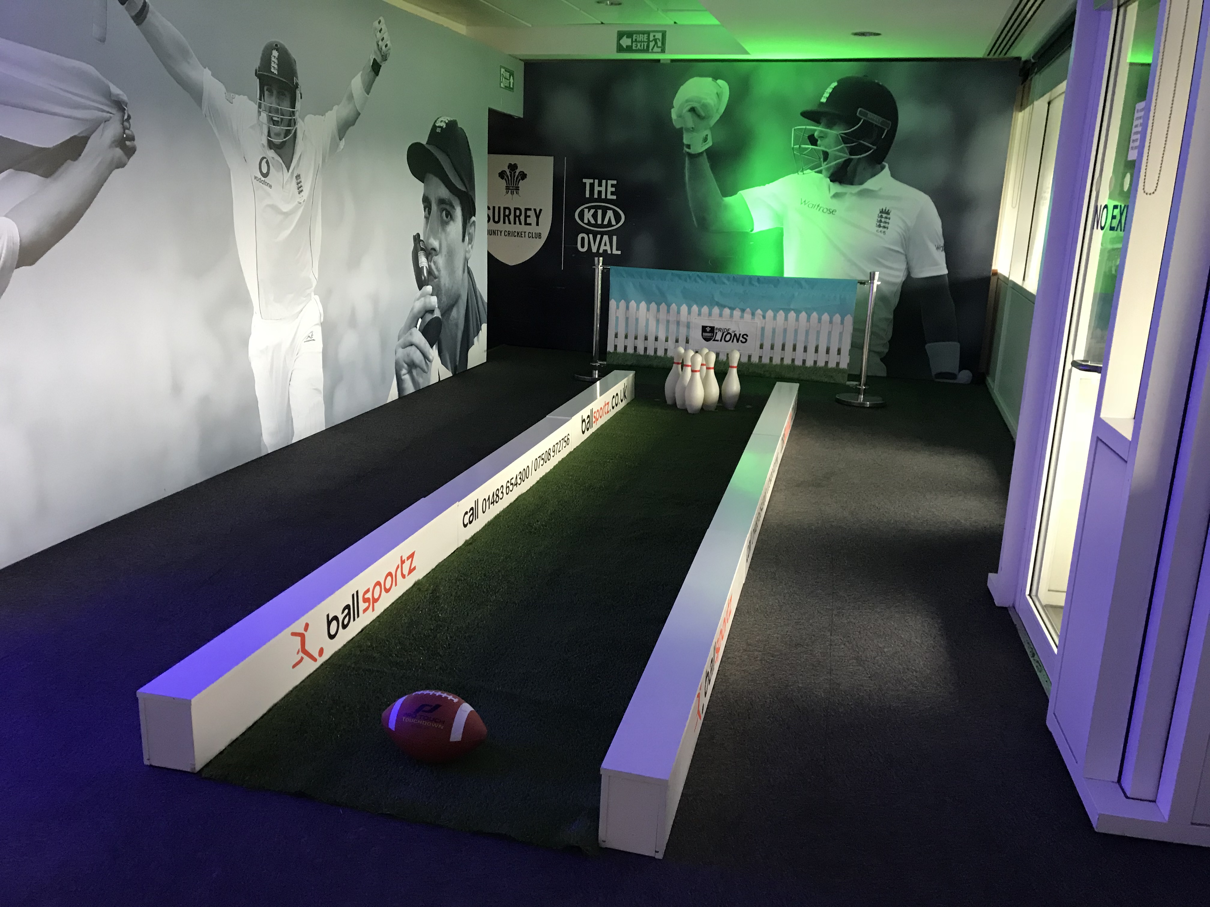 Indoor sports setup with bowling pins at the end of a narrow lane and an American football. Cricket-themed wall mural in the background.