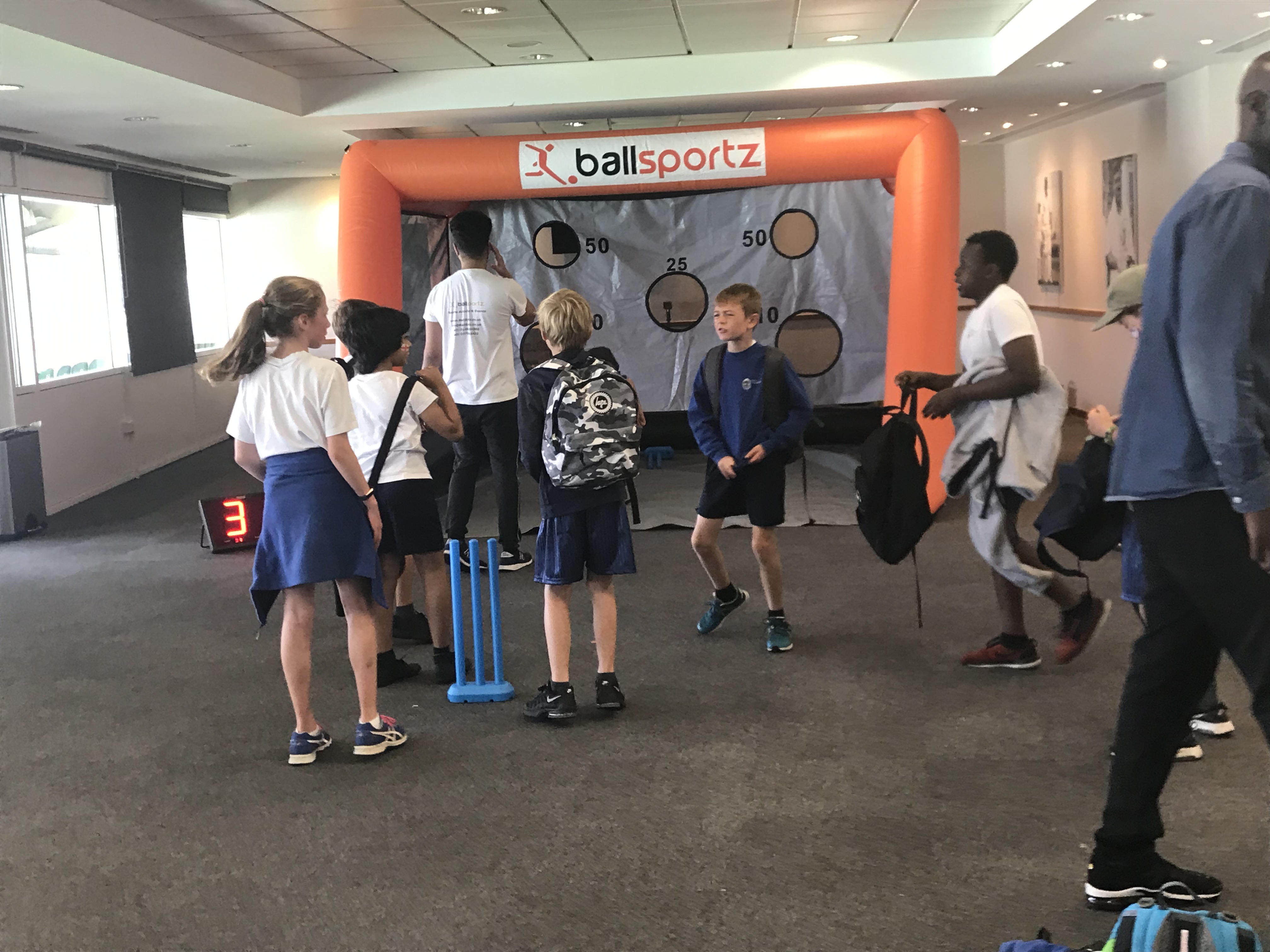 Children play a cricket game indoors in front of an inflatable target with numbers. A scoreboard reads 3. A person in a ballsportz shirt stands nearby.