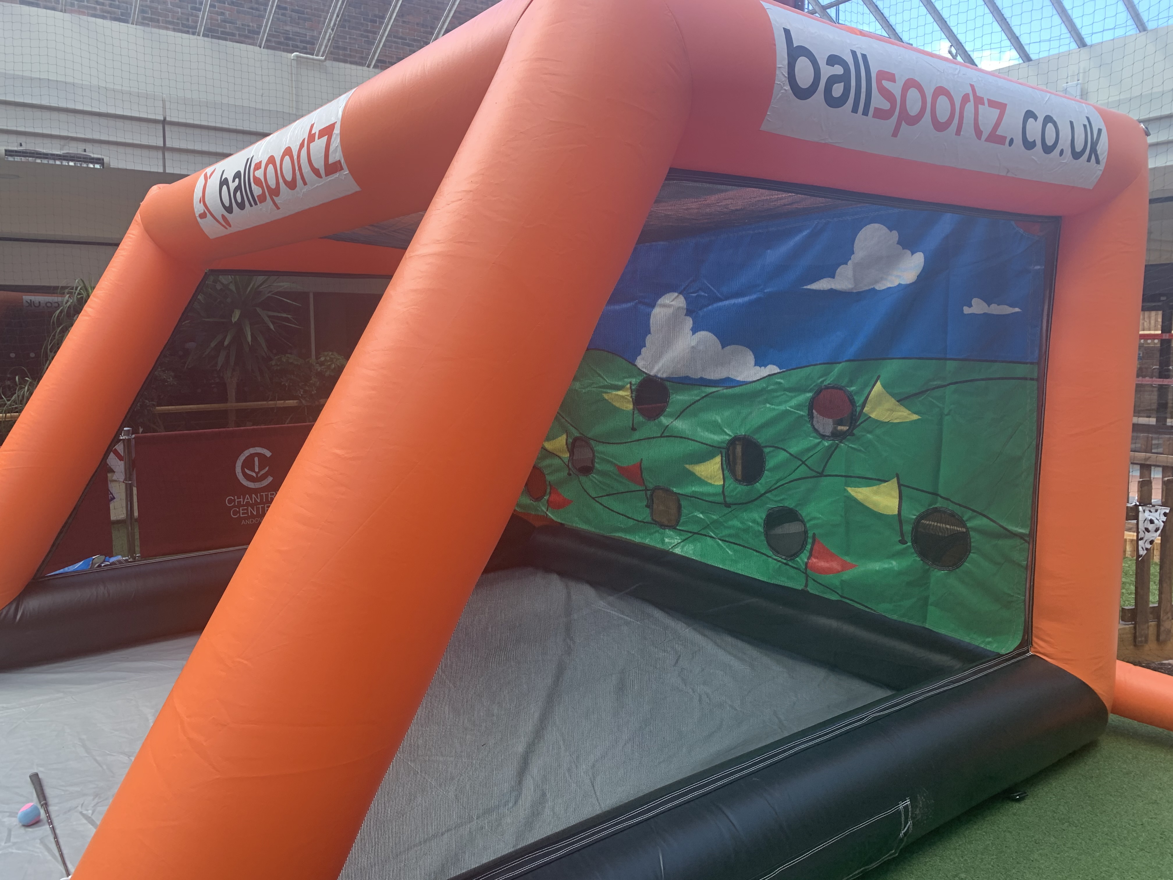 Inflatable, orange game booth with "ballsportz.co.uk" branding. The netted area has a backdrop of a colorful landscape with trees and a sky.