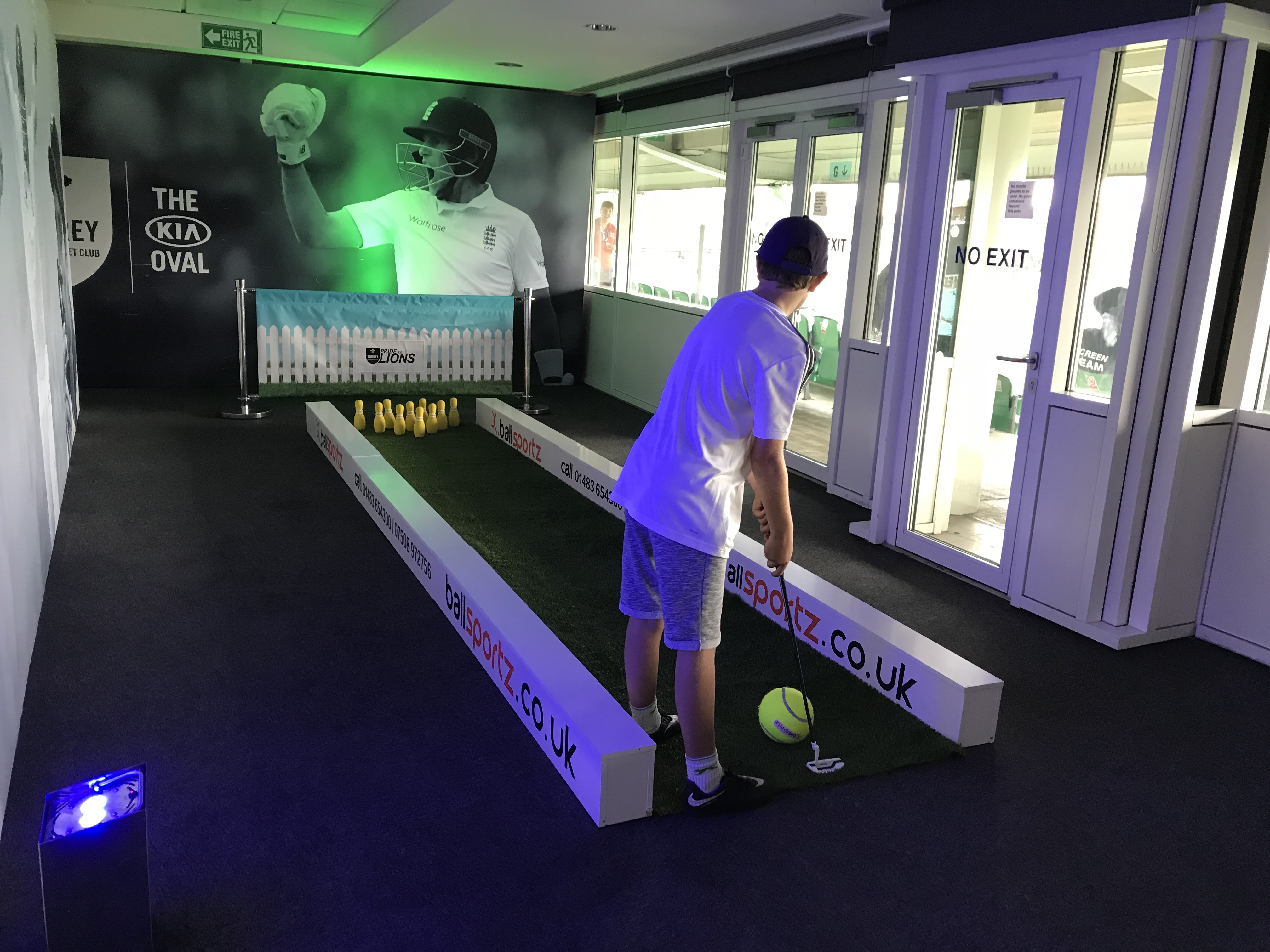 A person plays mini-golf indoors, aiming at bowling pins in a narrow, turf-covered lane.