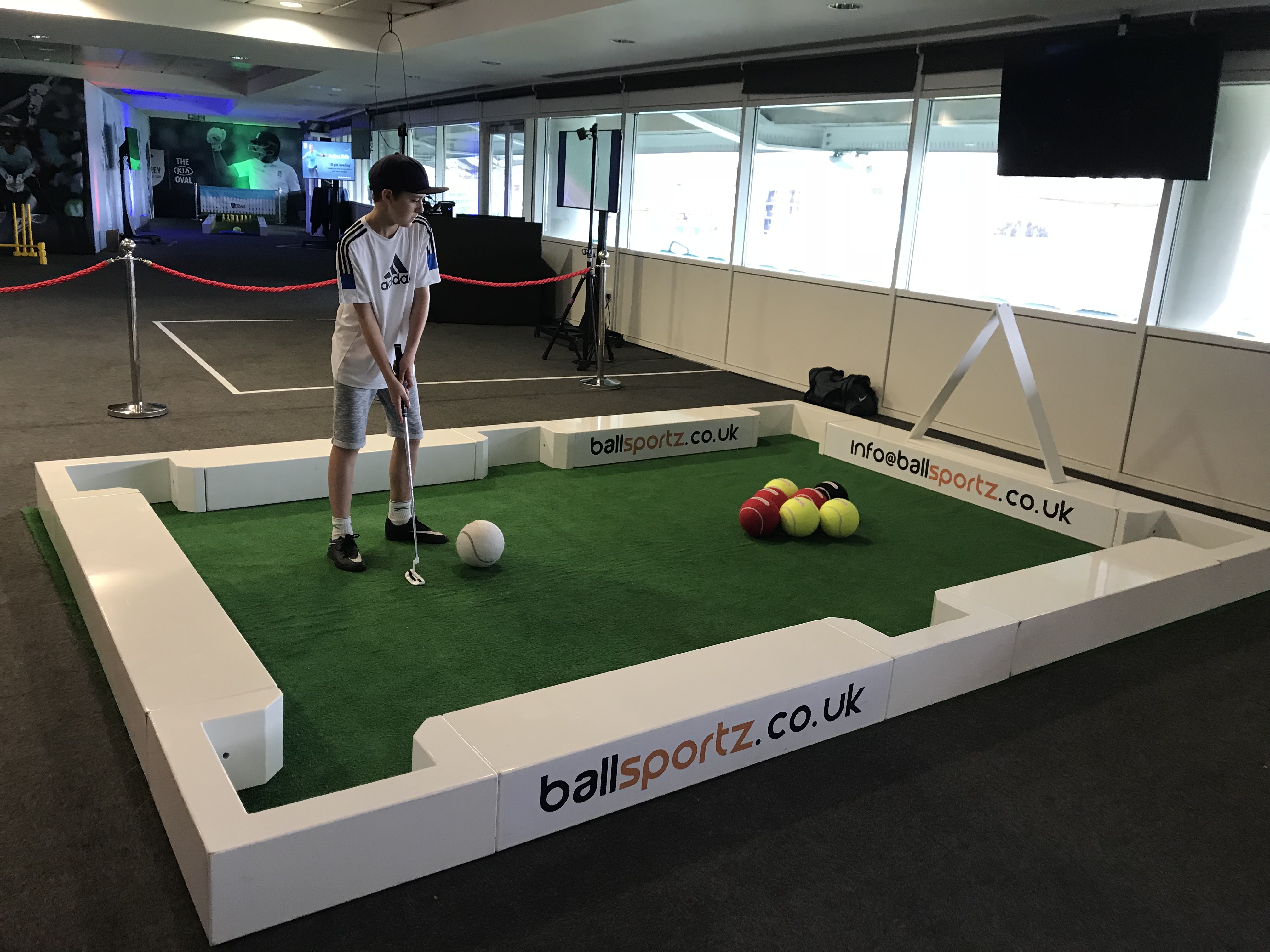 A person playing a hybrid game of football and pool, using a cue to hit a large white ball towards colored balls in an enclosed area.