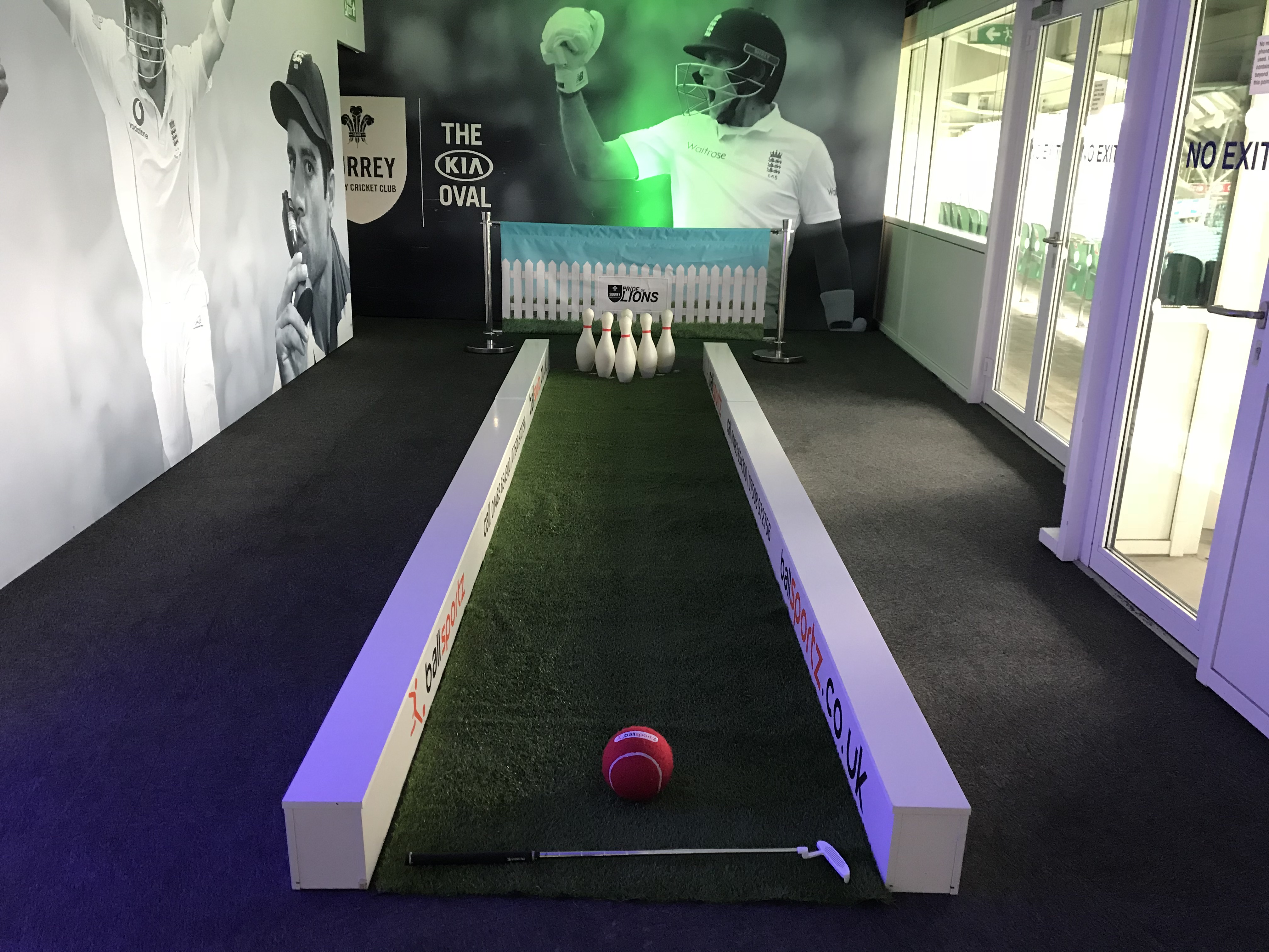 Indoor cricket-themed bowling alley with a red cricket ball, white pins, and a small bat on green turf, surrounded by cricket player images.
