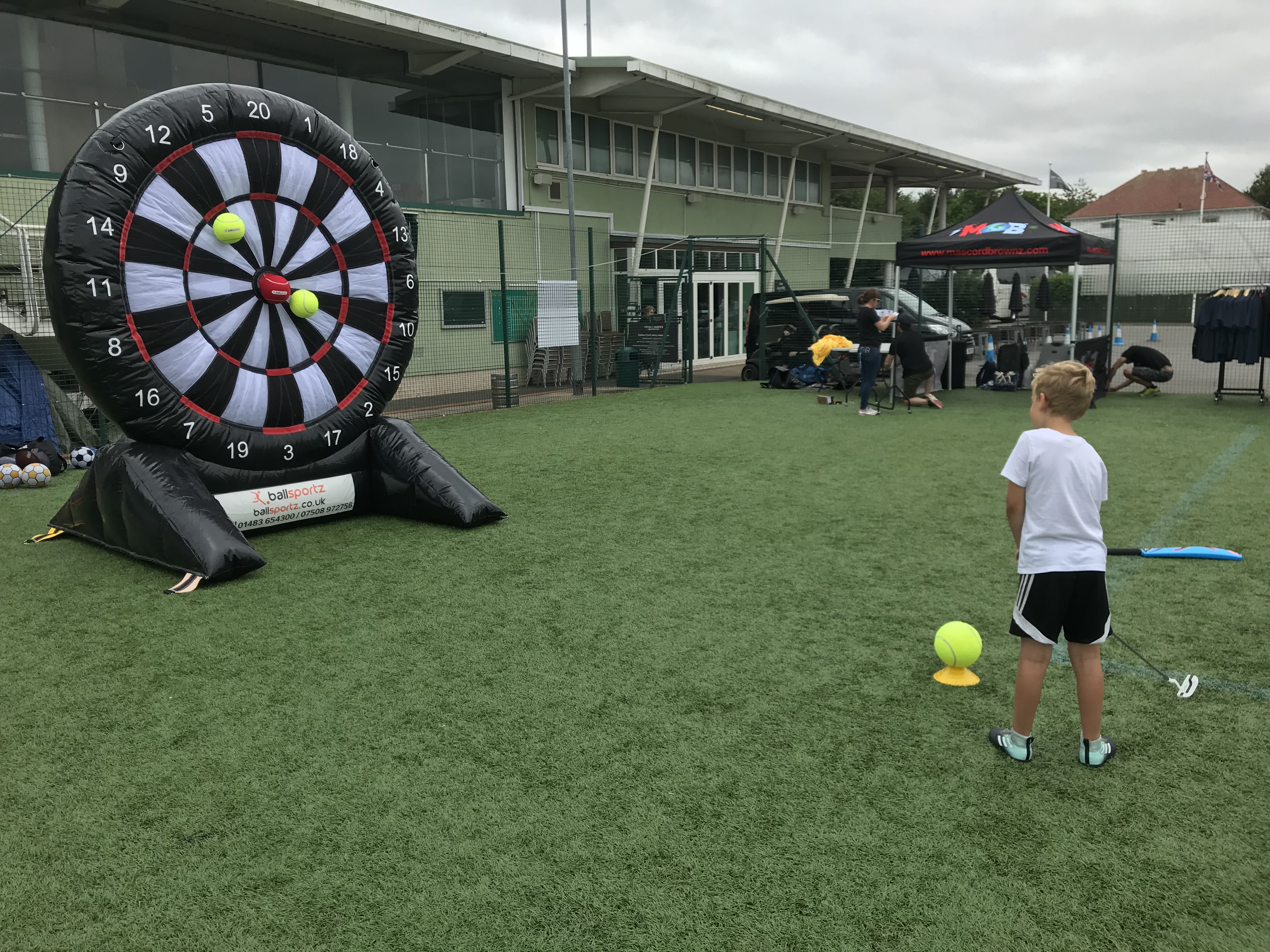 A child aims a soccer ball at a large inflatable dartboard on a sports field. Spectators and various equipment are visible in the background.