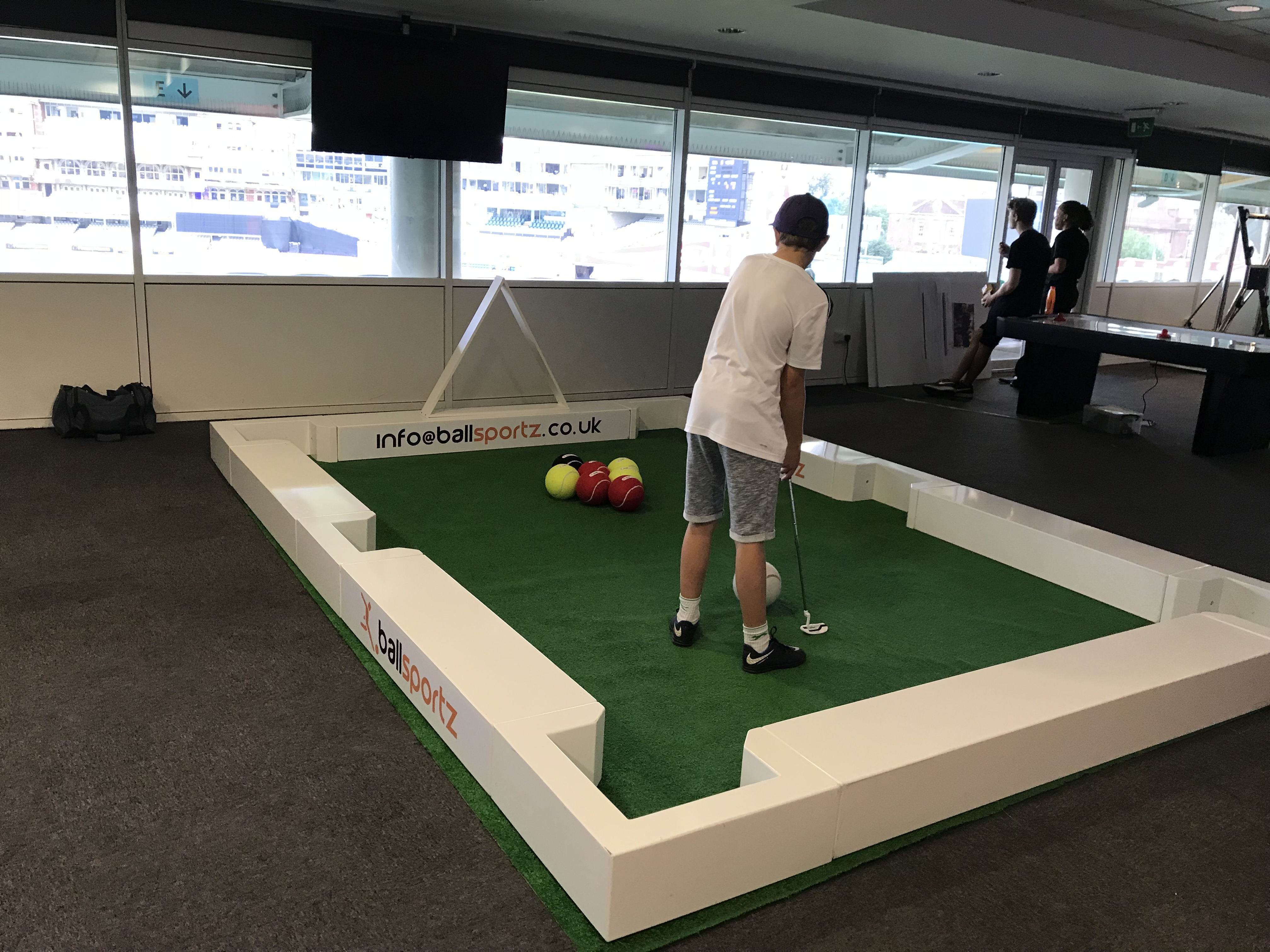 Person playing mini-golf on an indoor artificial grass course with colorful balls and white barriers.