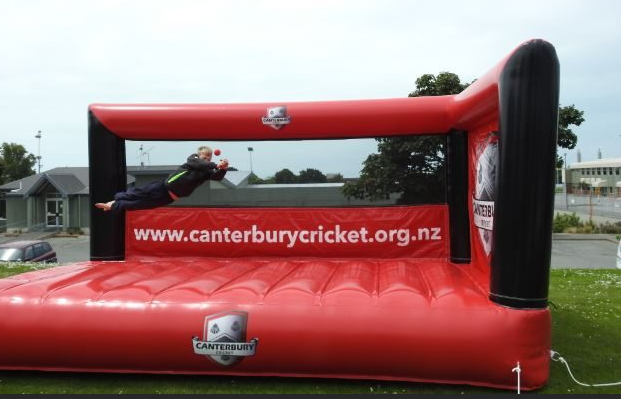 Person diving onto a red inflatable structure with the website "canterburycricket.org.nz" printed on it, outdoors on a cloudy day.