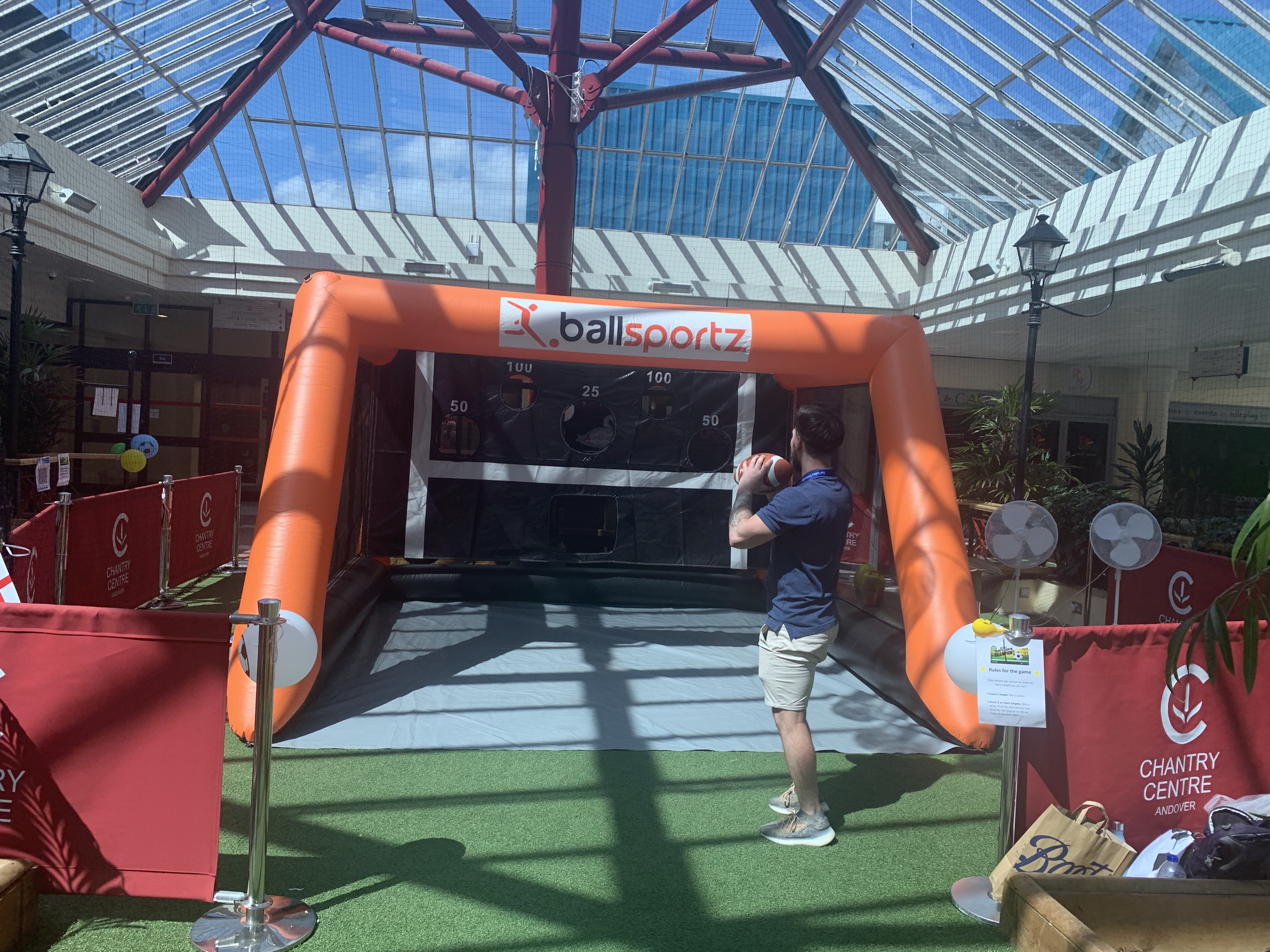 Person playing a ball toss game inside an inflatable booth labeled "ballsportz" in a sunlit atrium.