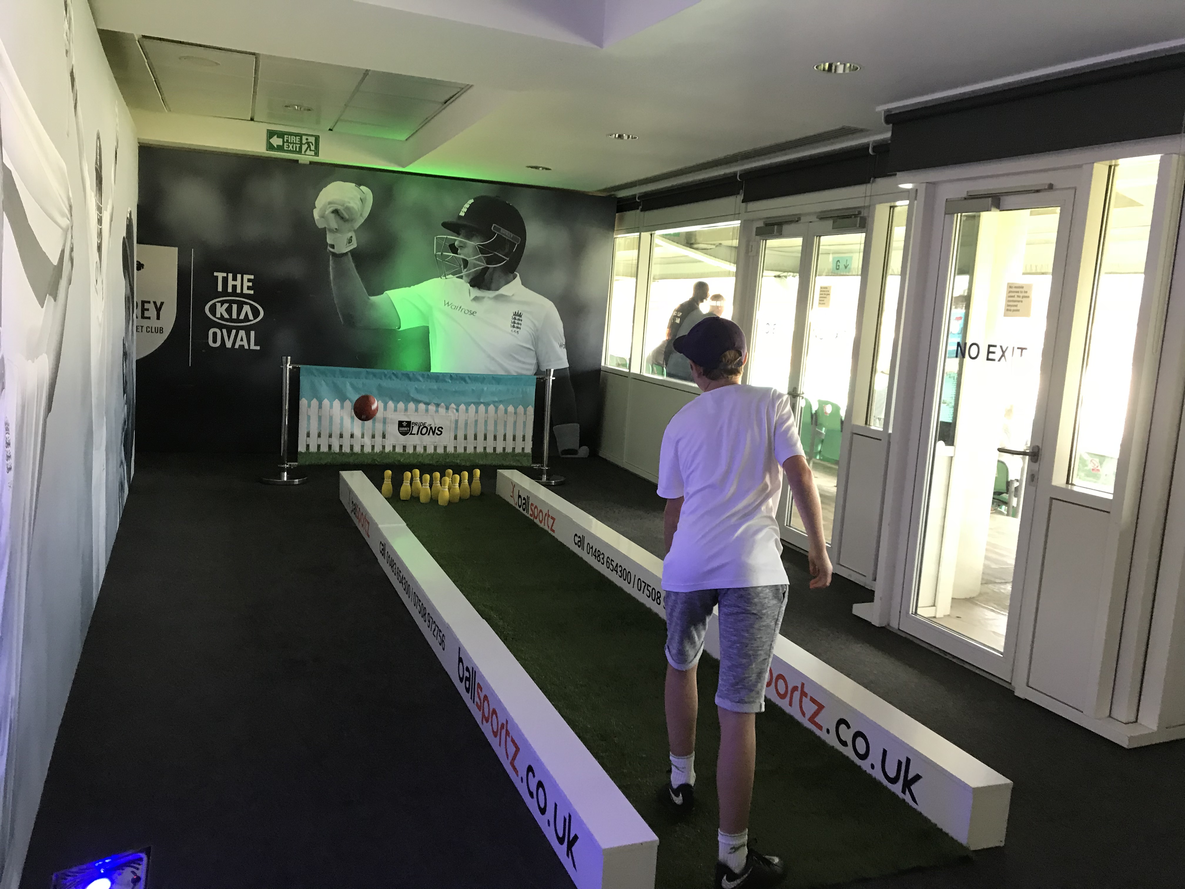 Child plays a bowling game in an indoor cricket-themed area with promotional banners.