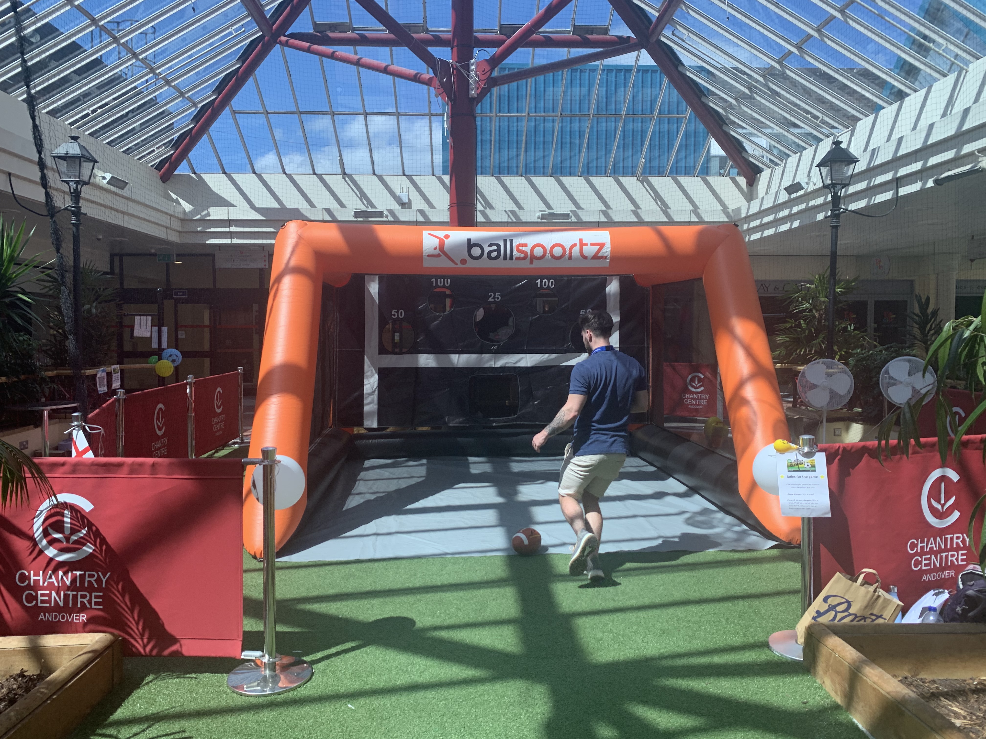 A person kicks a soccer ball into an inflatable goal at an indoor sports setup with a clear roof. Chantry Centre signage is visible.