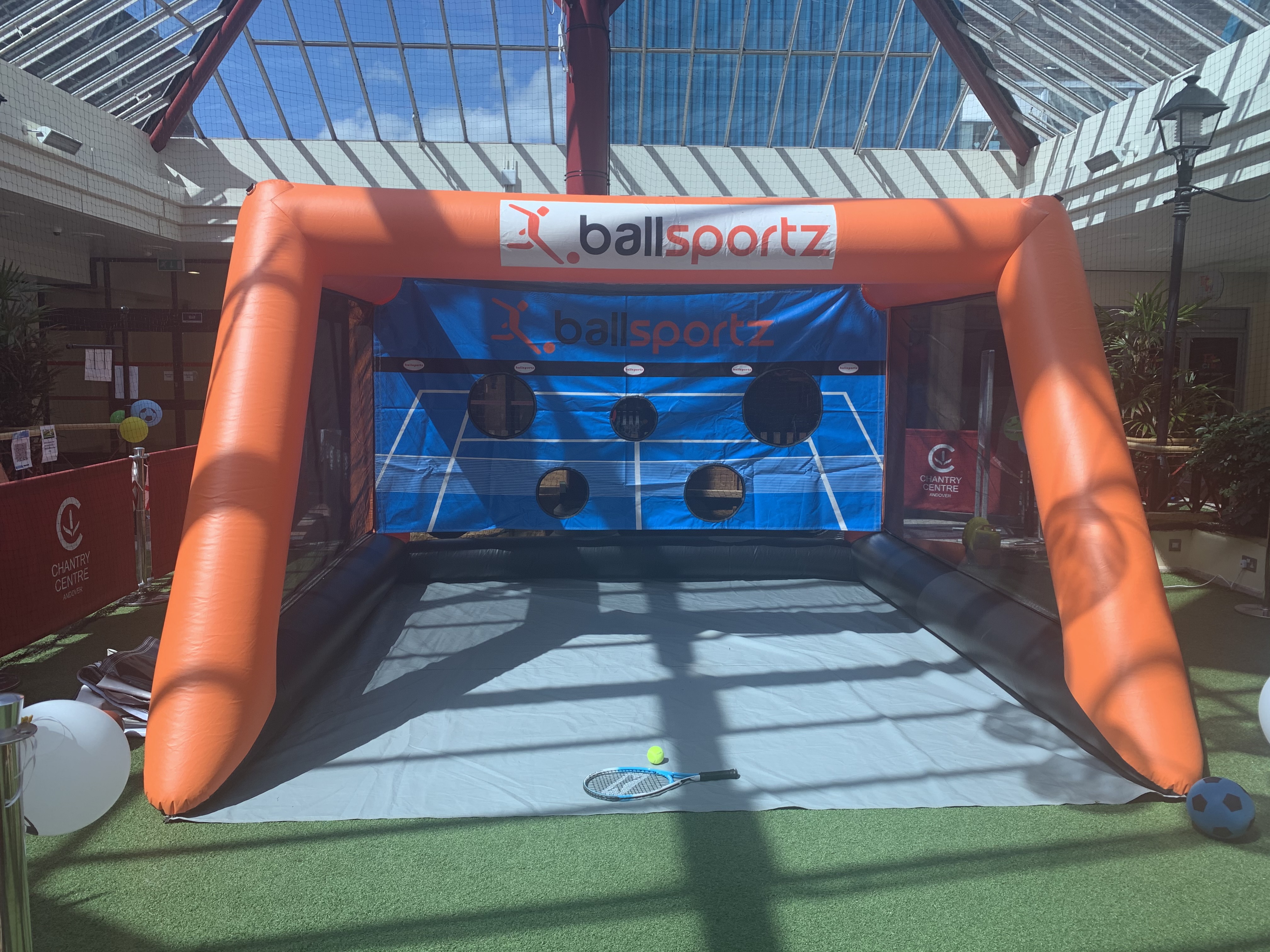 An inflatable soccer goal setup with targets in a sunny indoor area. A tennis racket lies on the ground in front.