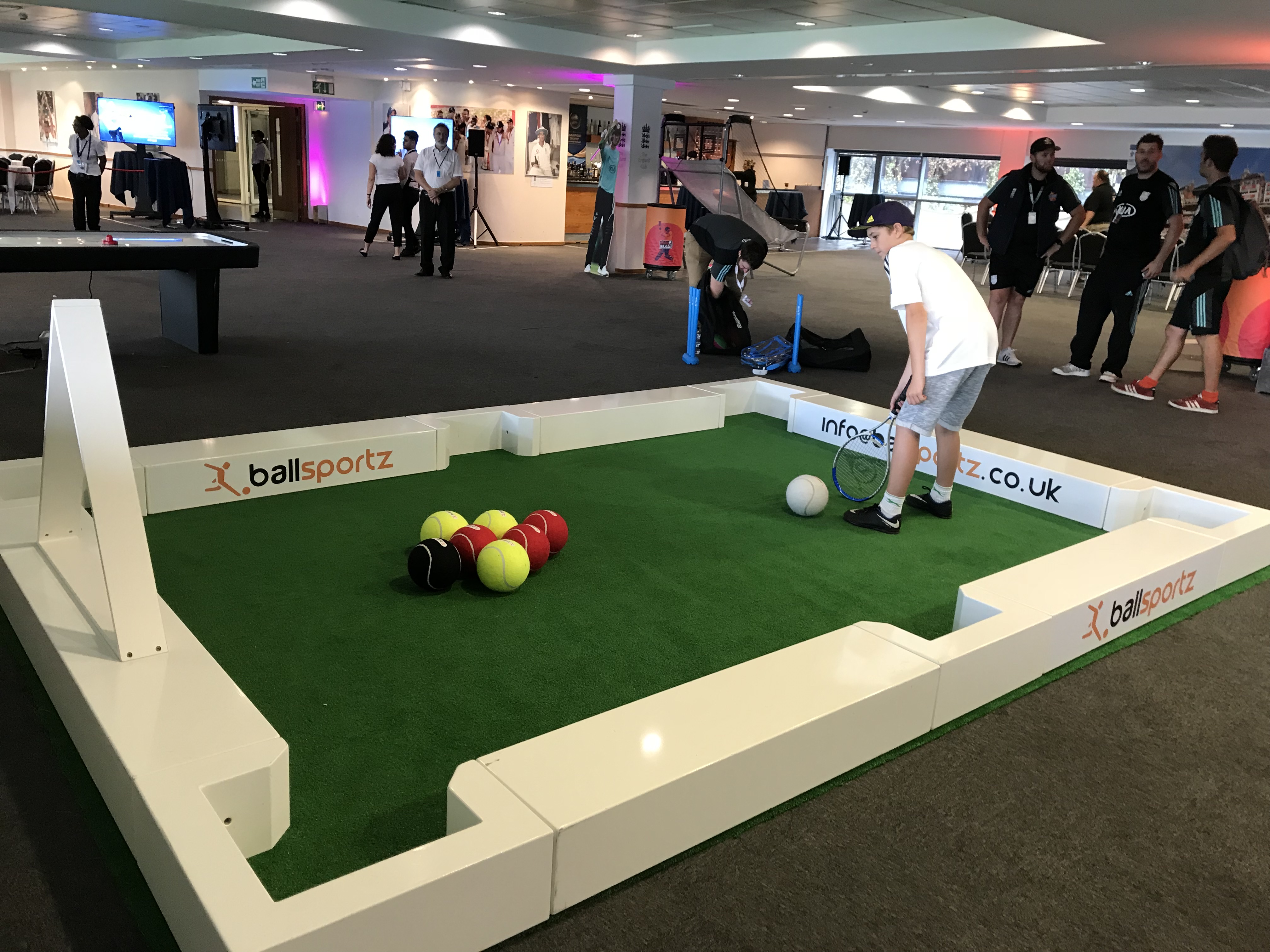 Person playing a hybrid game of pool and soccer using colorful balls on a large green carpet. Others are observing in the background.
