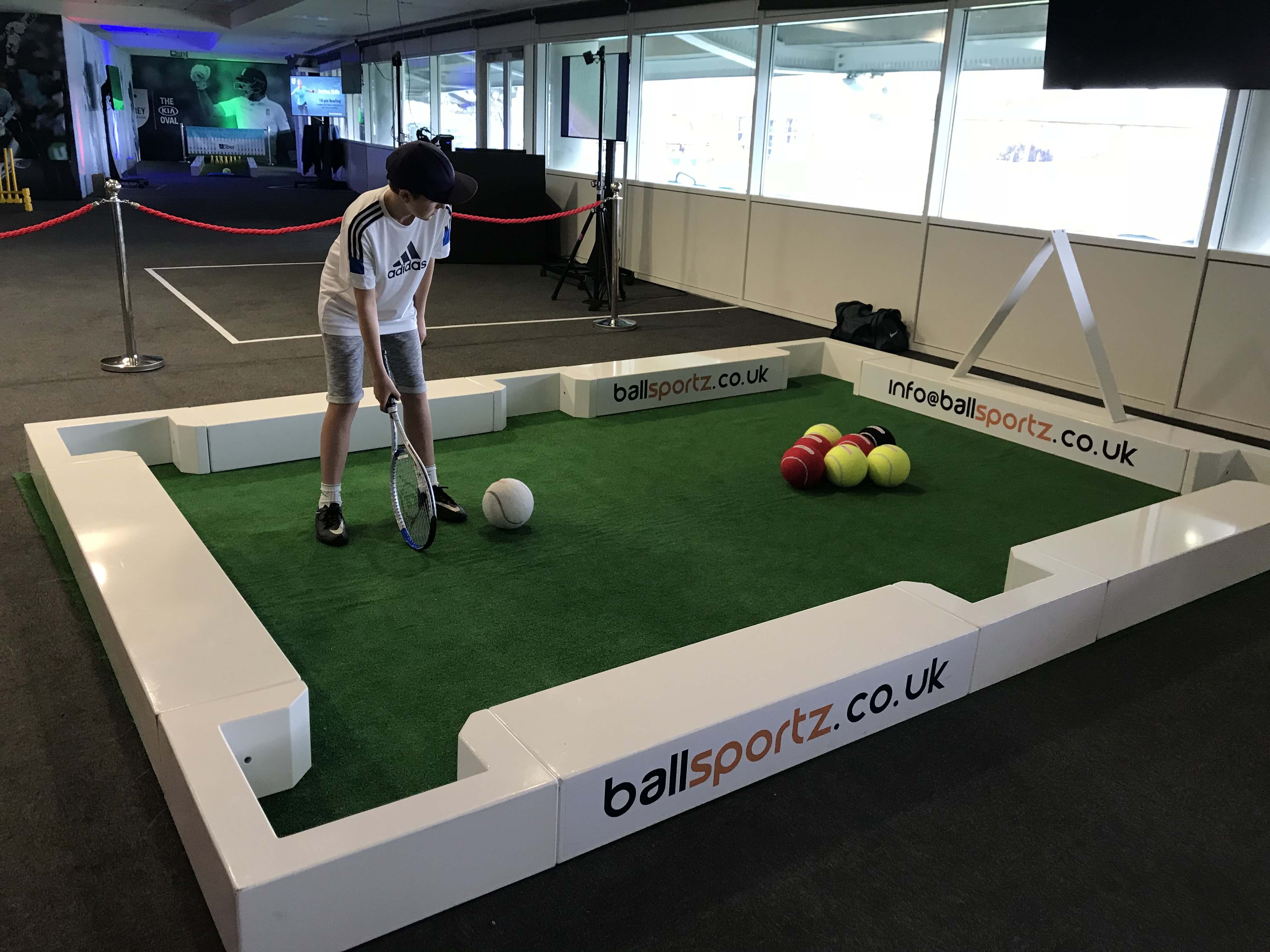 A person in sports attire plays a game on a miniature artificial turf court, using a tennis racket to hit a large ball toward smaller balls. The court is branded with “ballsportz.co.uk.”.