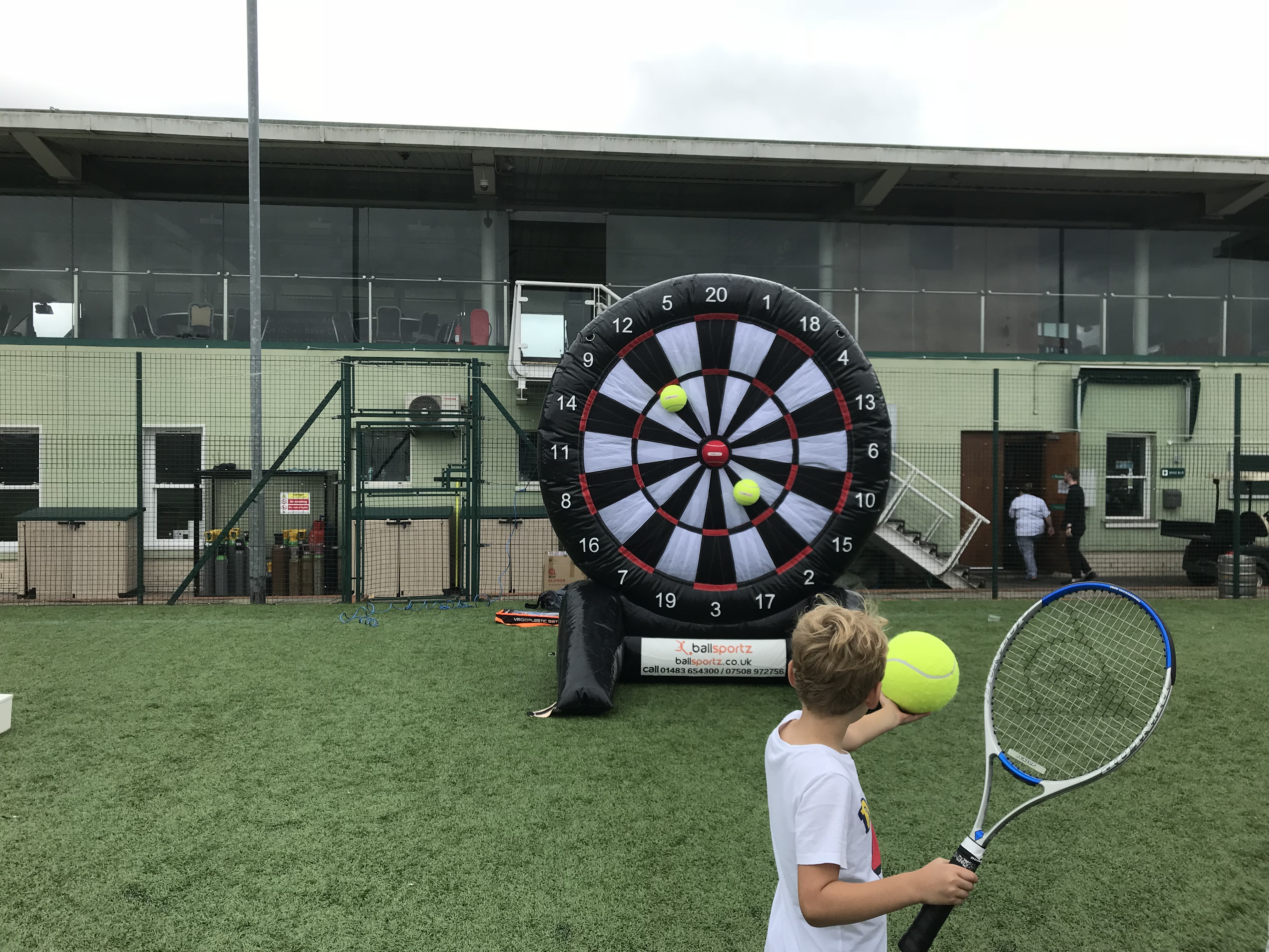 Child holding a tennis racket stands in front of a large inflatable dartboard with tennis balls attached.
