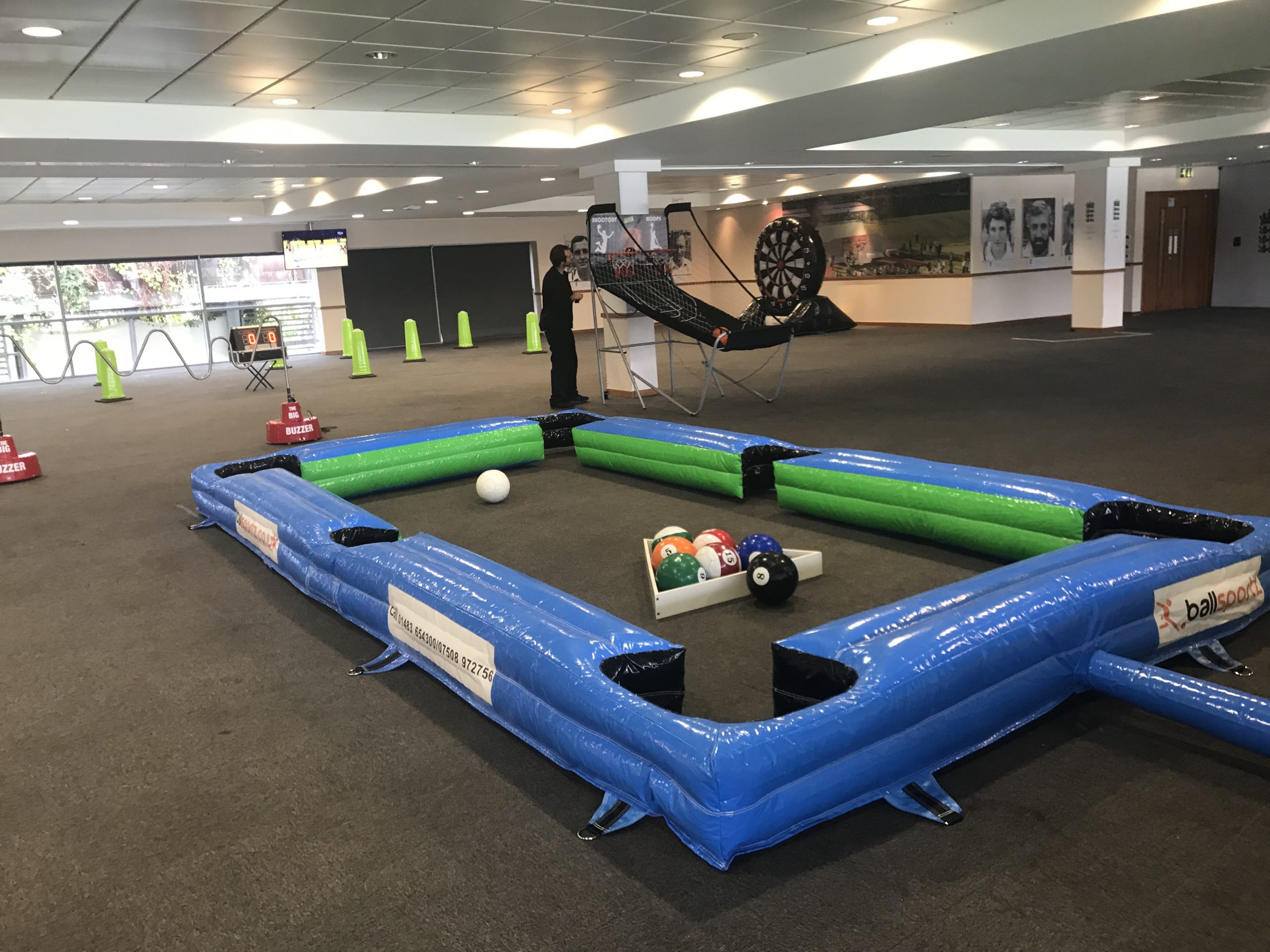 Indoor inflatable soccer billiards game setup with colorful balls, enclosed by blue and green barriers, and a nearby person standing.