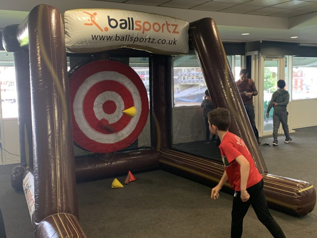 A child in a red shirt throws a dart at an inflatable target game indoors. Two others watch in the background.