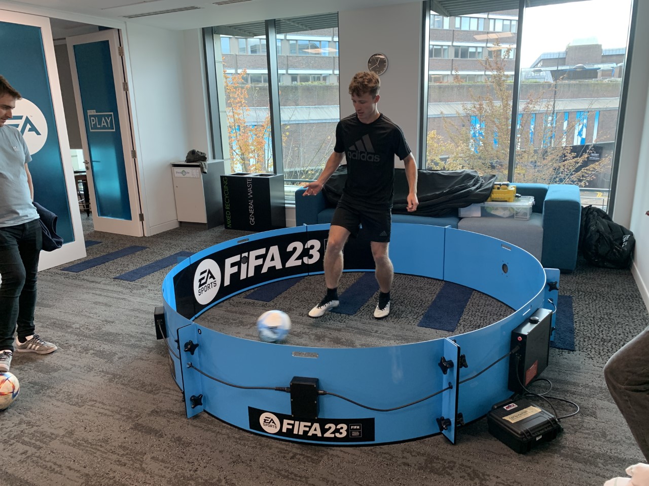 A person in sportswear plays football in a small, circular FIFA 23-themed arena indoors.
