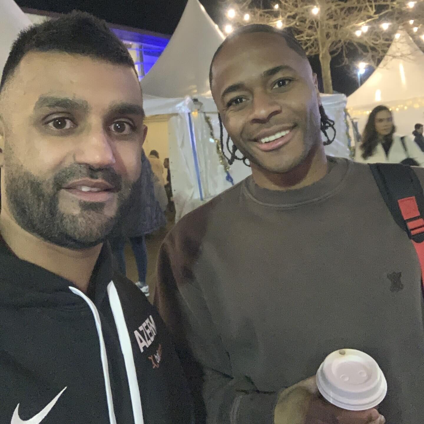 Two men pose for a selfie at an outdoor event, one holding a coffee cup. Tents and string lights are visible in the background.