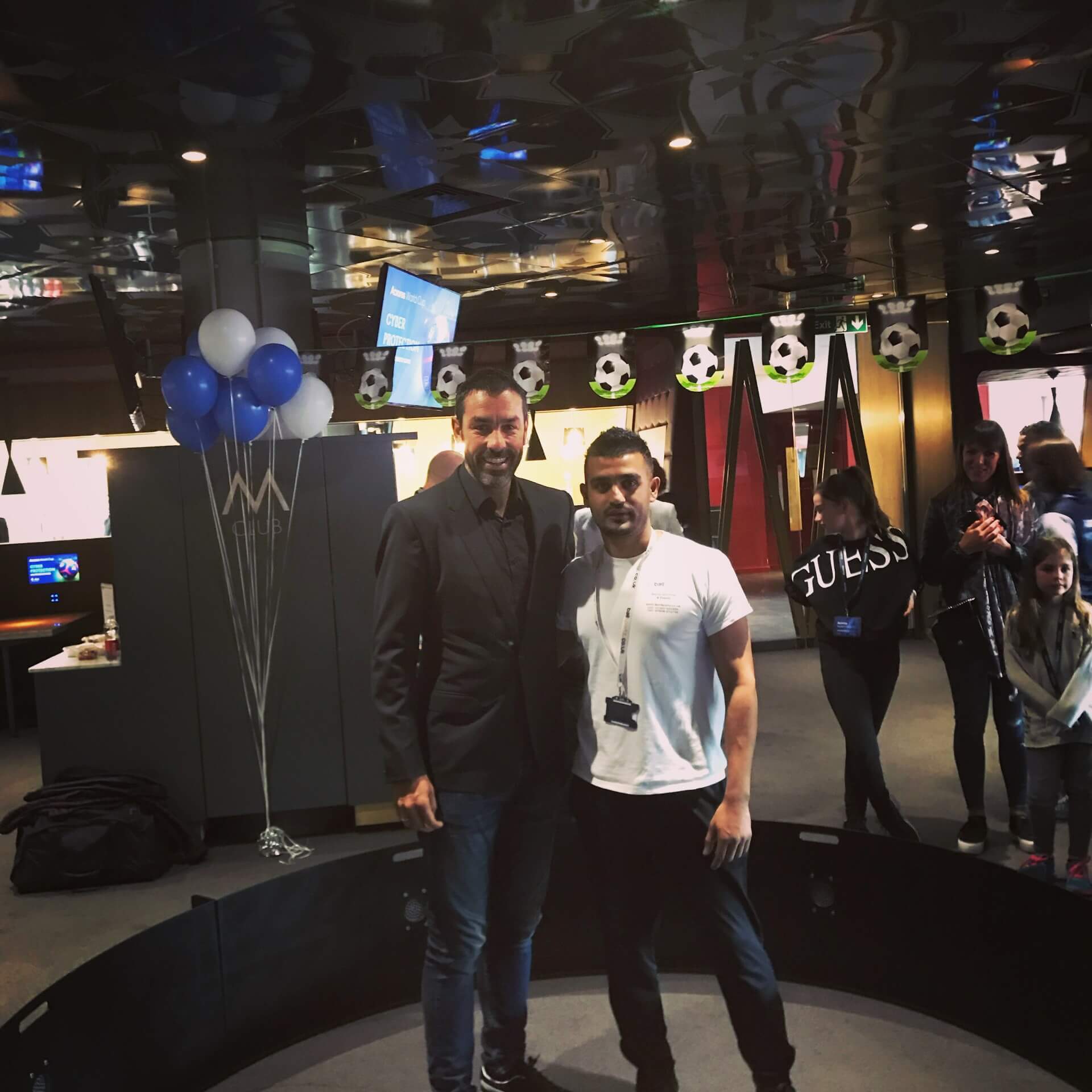 Two men standing together indoors, with balloons and soccer-themed decorations in the background.