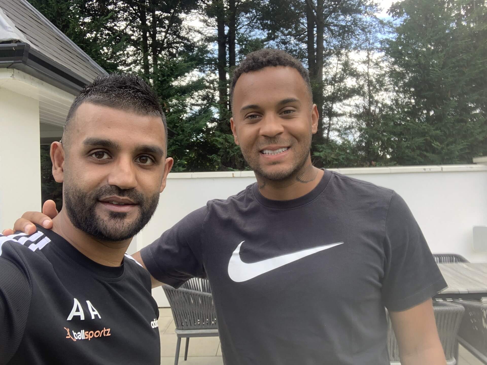 Two men standing outdoors, smiling at the camera. The man on the left wears a black shirt with "AAsports" text, while the man on the right wears a Nike T-shirt. Trees are visible in the background.