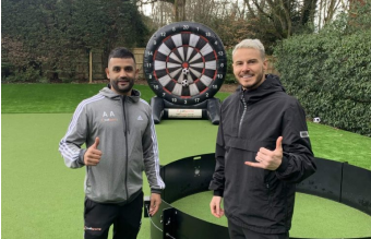 Two people posing in front of a giant dartboard outdoors, each making a gesture with one hand.
