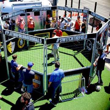People play soccer in a small, enclosed panna cage with spectators watching around. Banners with text are displayed on the structure.