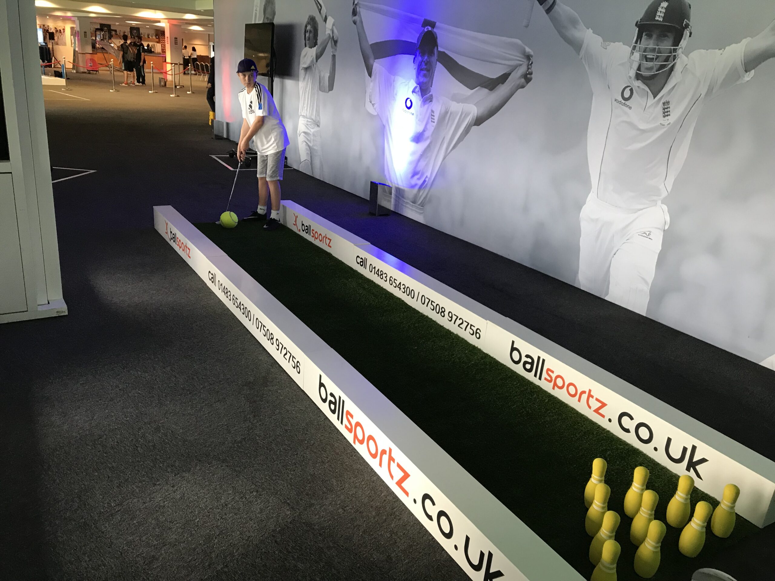 A person enjoys indoor mini-golf on a green turf adorned with giant images of cricketers. Bowling pins stand lined up as targets, offering a unique twist reminiscent of foot pool fun.