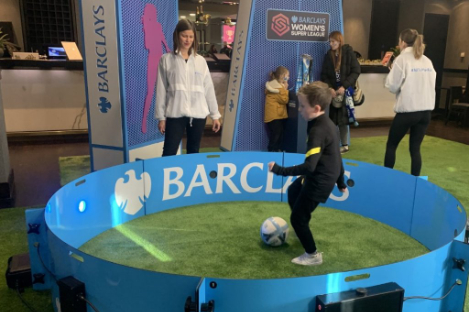 A child plays foot pool inside a circular blue enclosure with Barclays branding, while two adults observe.