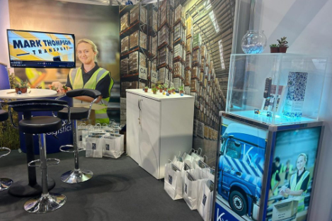 The trade show booth for "Mark Thompson Transport" features a captivating display, complete with promotional bags, a sleek glass case showcasing items, bar stools for comfort, and a TV screen showing a person in high-visibility clothing. A foot pool game adds an engaging touch to the setup.