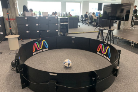 In an office space, a round indoor soccer playing area is cleverly set up like a foot pool. Amidst the playful setup, people diligently work in the background.