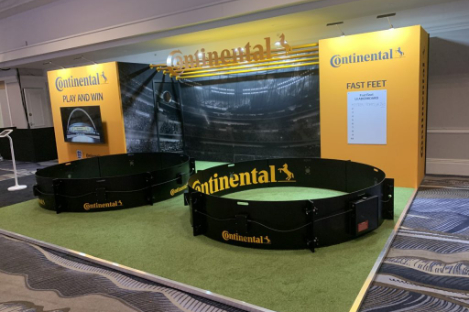 The exhibition booth, adorned with Continental branding, showcases an exciting foot pool setup within two small circular enclosures on vibrant green flooring. Banners boldly display "Play and Win" and "Fast Feet," enticing visitors to join the fun and test their agility.