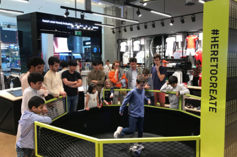 Children and adults watch a small soccer game, akin to a foot pool match, in a retail store surrounded by a fence adorned with the slogan "#HERETOCREATE.