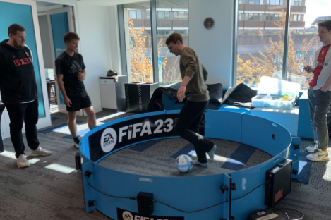Four people are gathered around a small circular soccer pitch labeled FIFA 23, reminiscent of a lively foot pool game. One person is skillfully playing with a soccer ball inside the pitch.