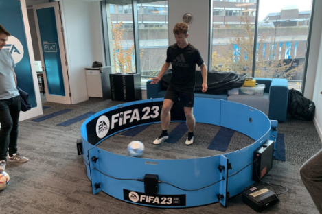 A person is skillfully playing foot pool with a soccer ball in a small blue circular arena labeled "FIFA 23" within an indoor setting.
