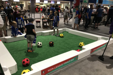 Children engage in a lively game of foot pool, kicking colored balls into pockets on a mini field at a bustling indoor event.