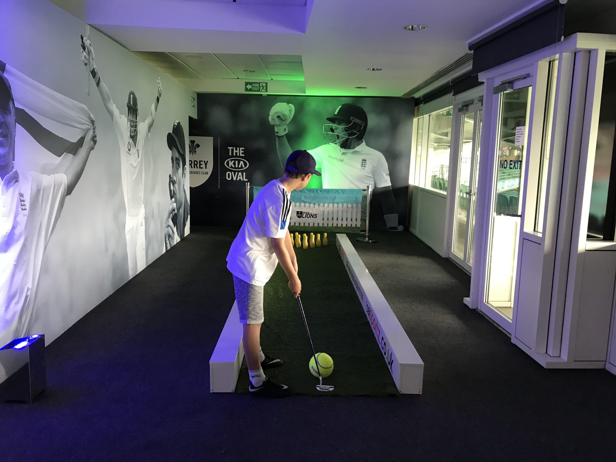 A person plays an indoor cricket bowling game, aiming at yellow pins. The room is decorated with large black-and-white cricket images.