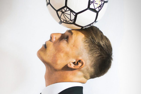 A person in a suit skillfully balances a soccer ball on their head, evoking the playful spirit of foot pool, all set against a crisp white background.