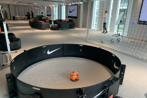 The room features a small circular soccer arena with an orange soccer ball and a goalpost in the background. Chairs, tables, and multiple TV screens displaying the Nike logo surround the setup, giving it a playful foot pool vibe.